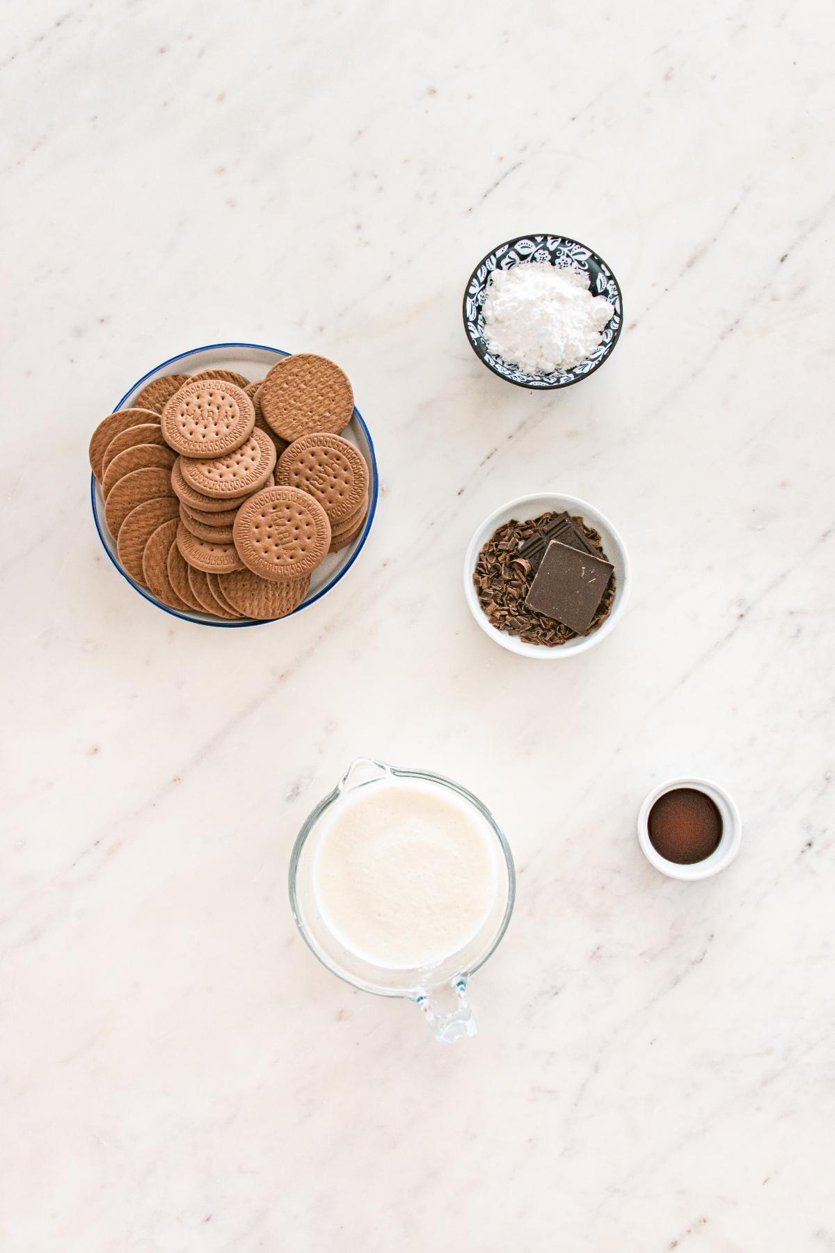 Biscuits, chocolate pieces, cream, cocoa powder, and powdered sugar arranged on a white marble surface.