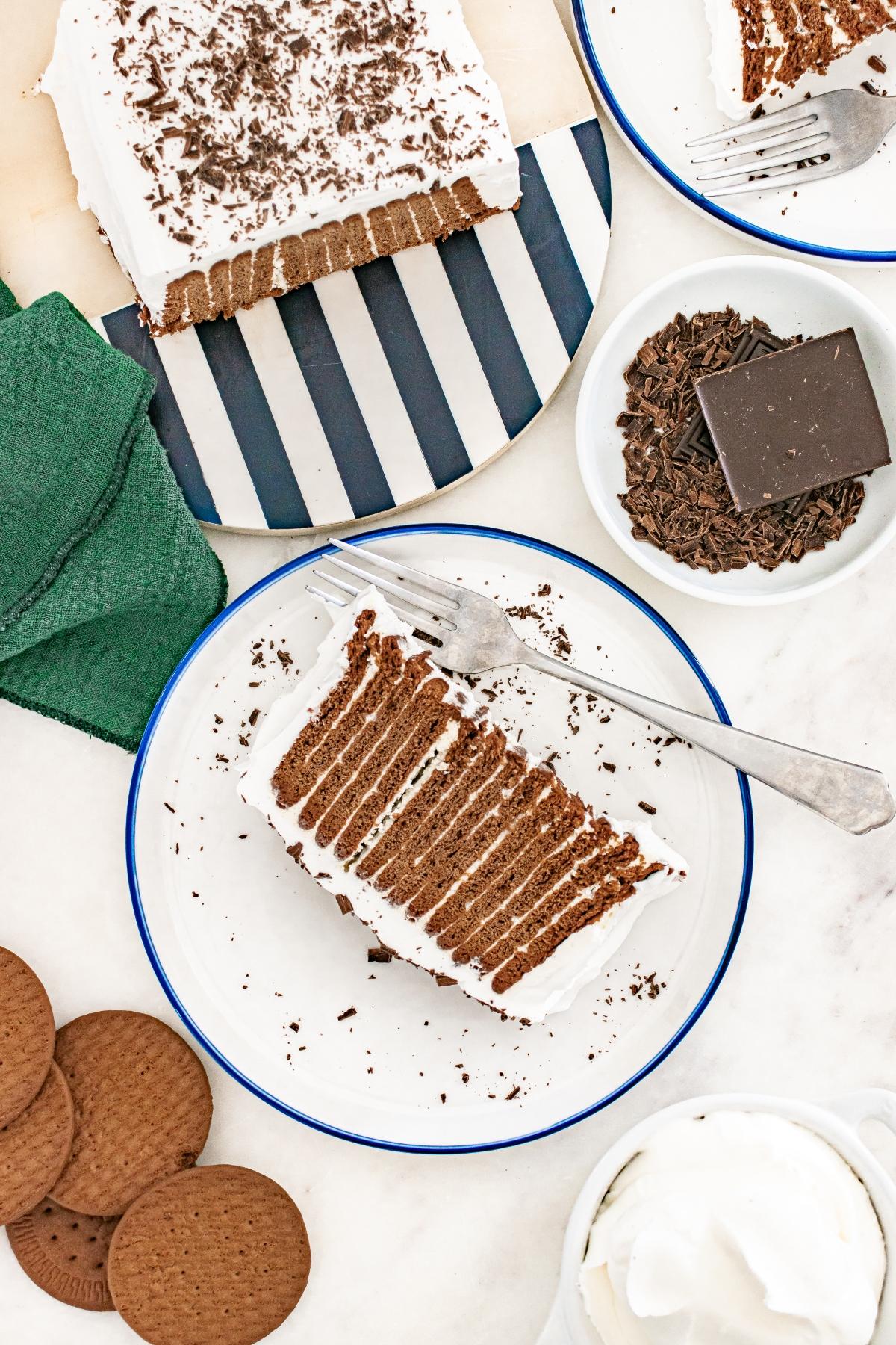 A slice of chocolate icebox cake with whipped cream on a plate, surrounded by cookies and chocolate shavings.