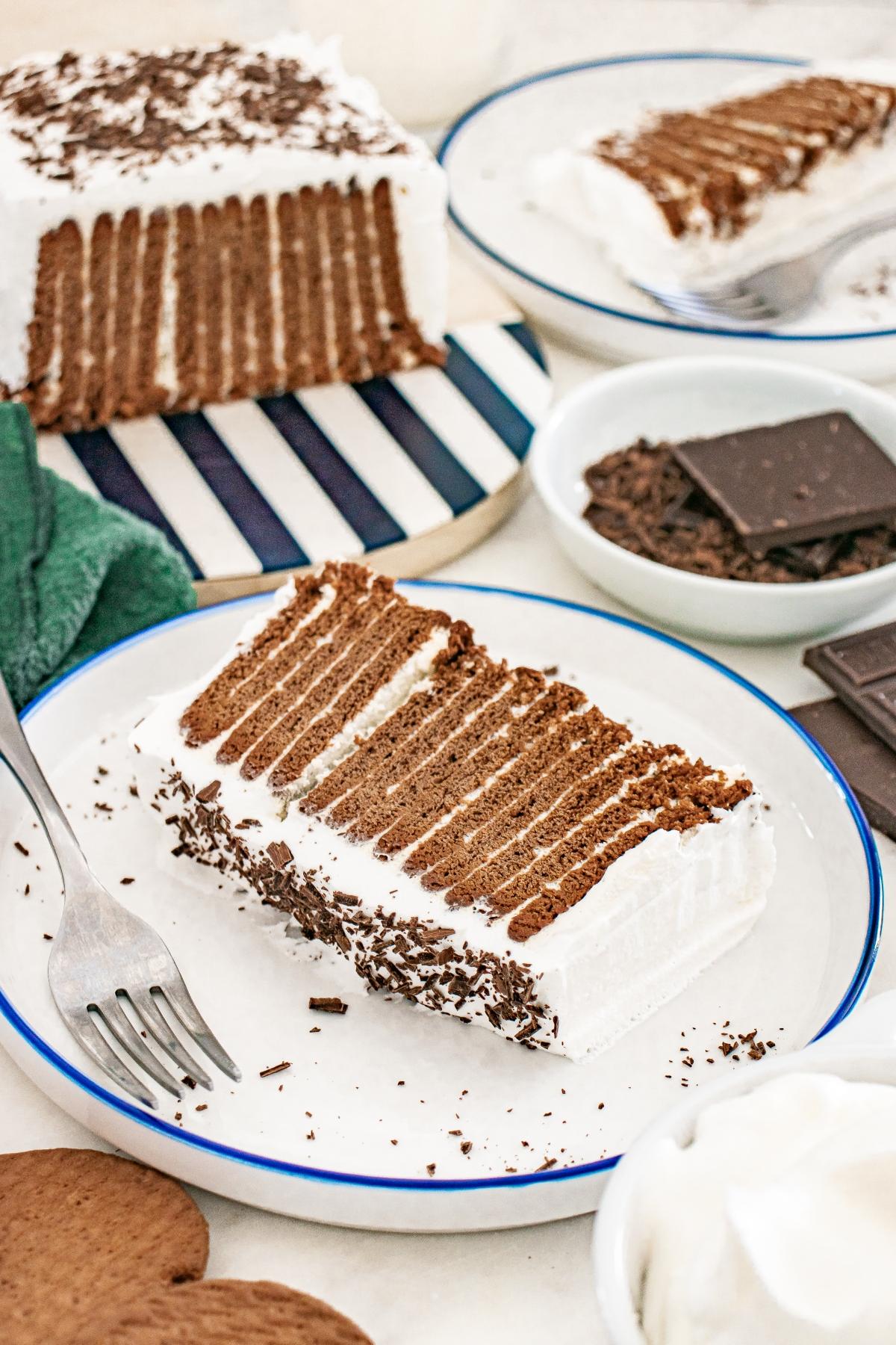 A slice of layered Chocolate icebox cake on a plate with a fork, surrounded by chocolate shavings.