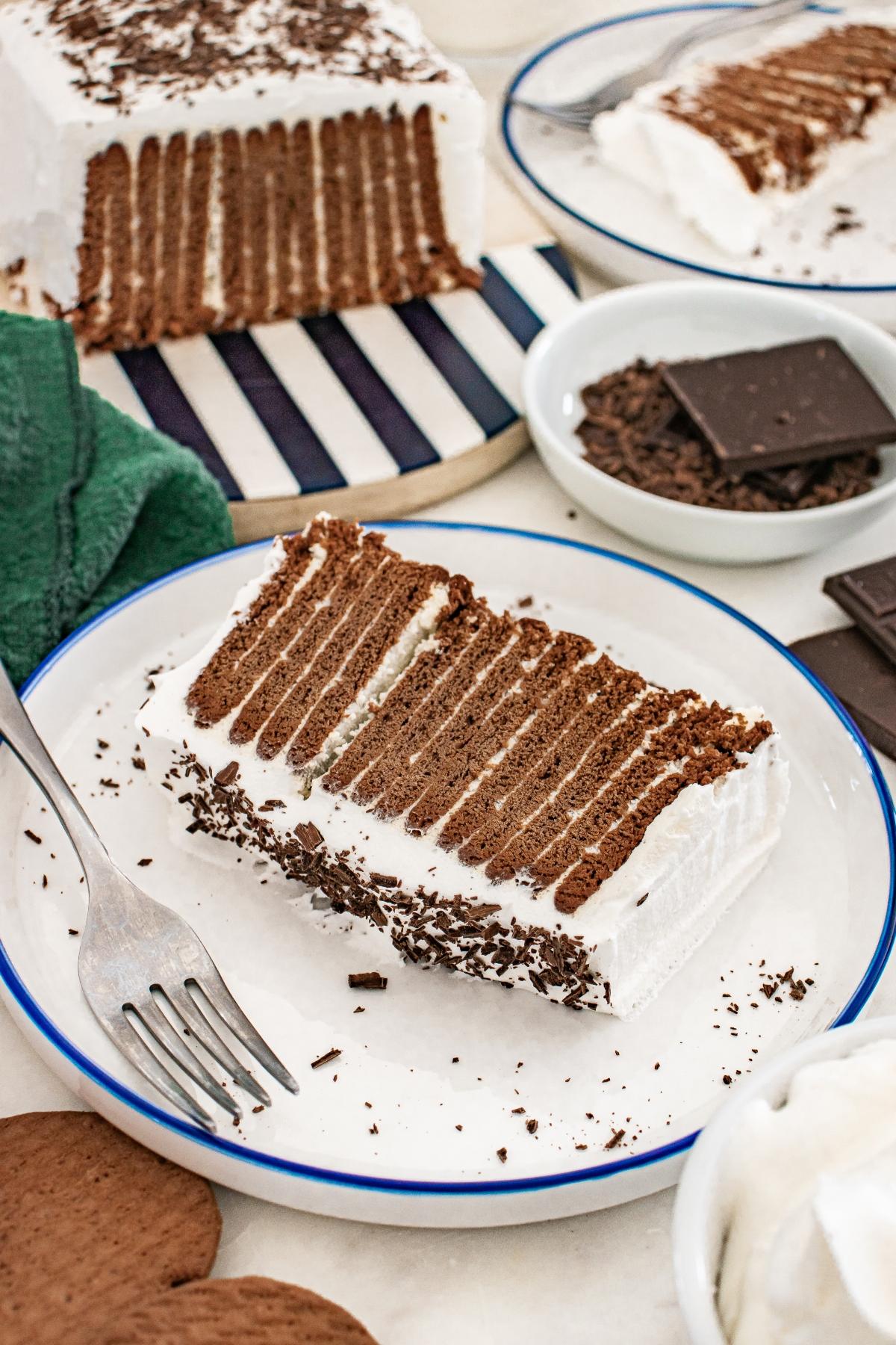 A slice of chocolate icebox cake with whipped cream and chocolate shavings on a white plate with a fork.