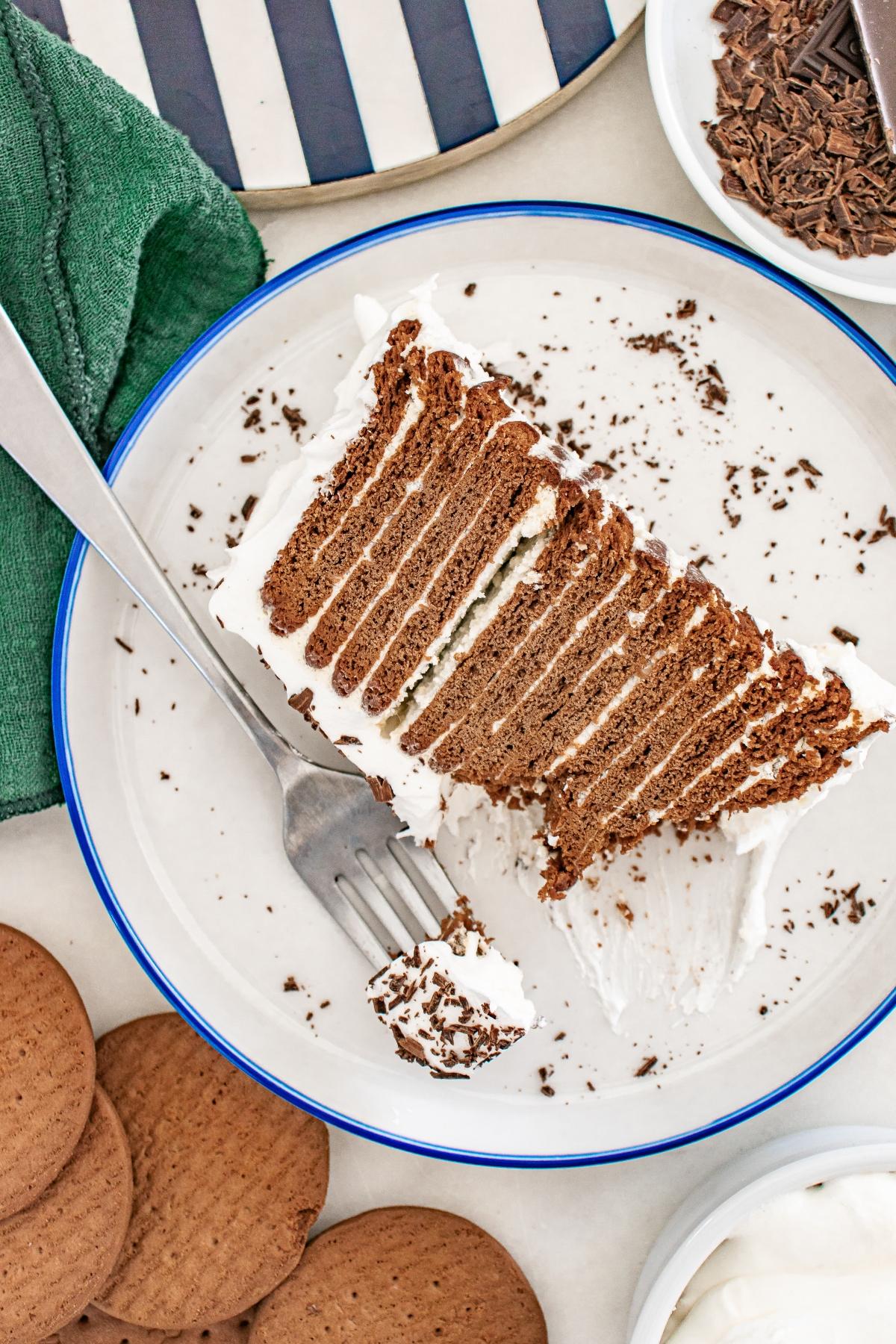 A slice of layered chocolate and cream cake on a white plate with a fork, surrounded by cookies and chocolate shavings.