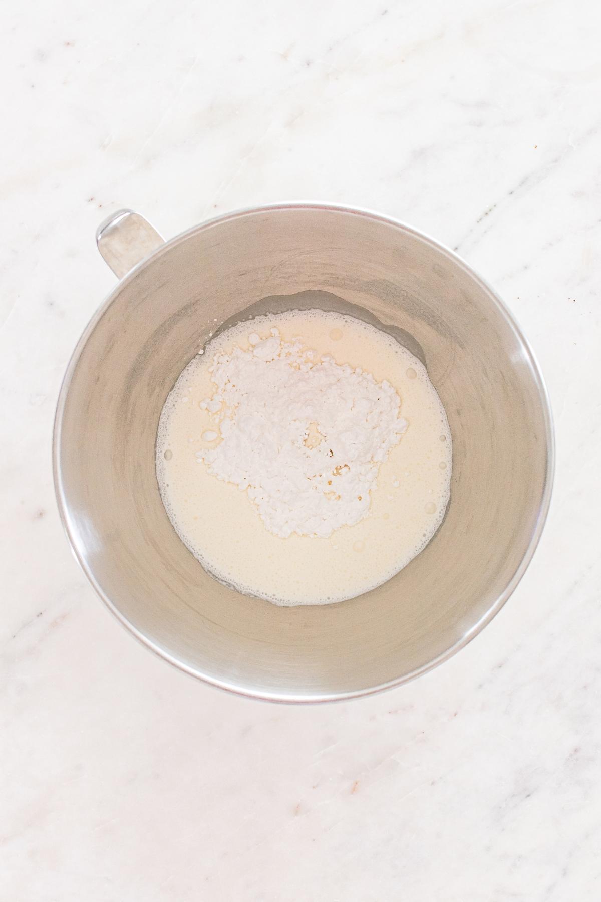 A metal mixing bowl with flour and yeast in liquid on a white marble surface.