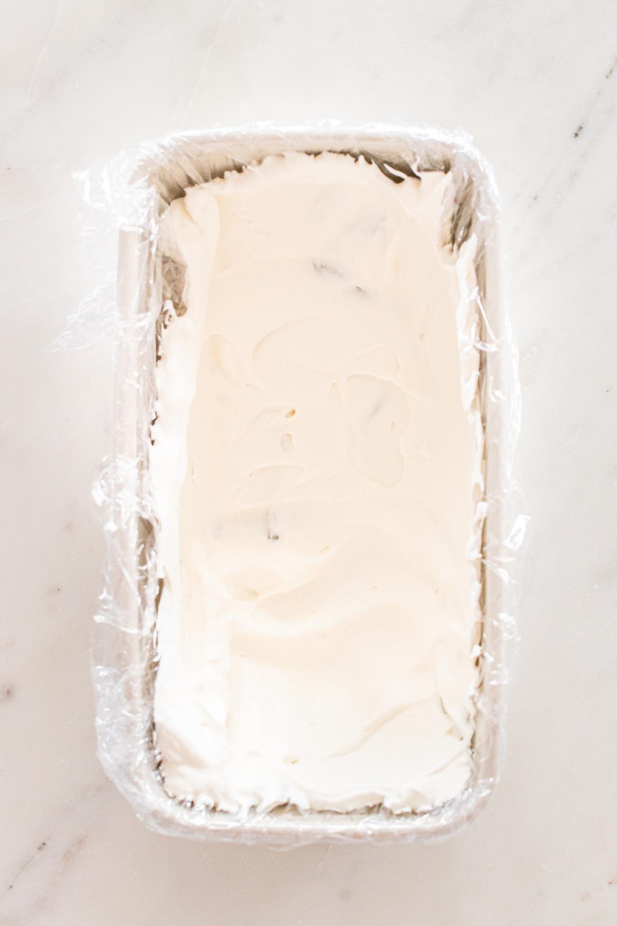 A loaf pan lined with plastic wrap filled with smooth, white whipped cream on a marble surface.