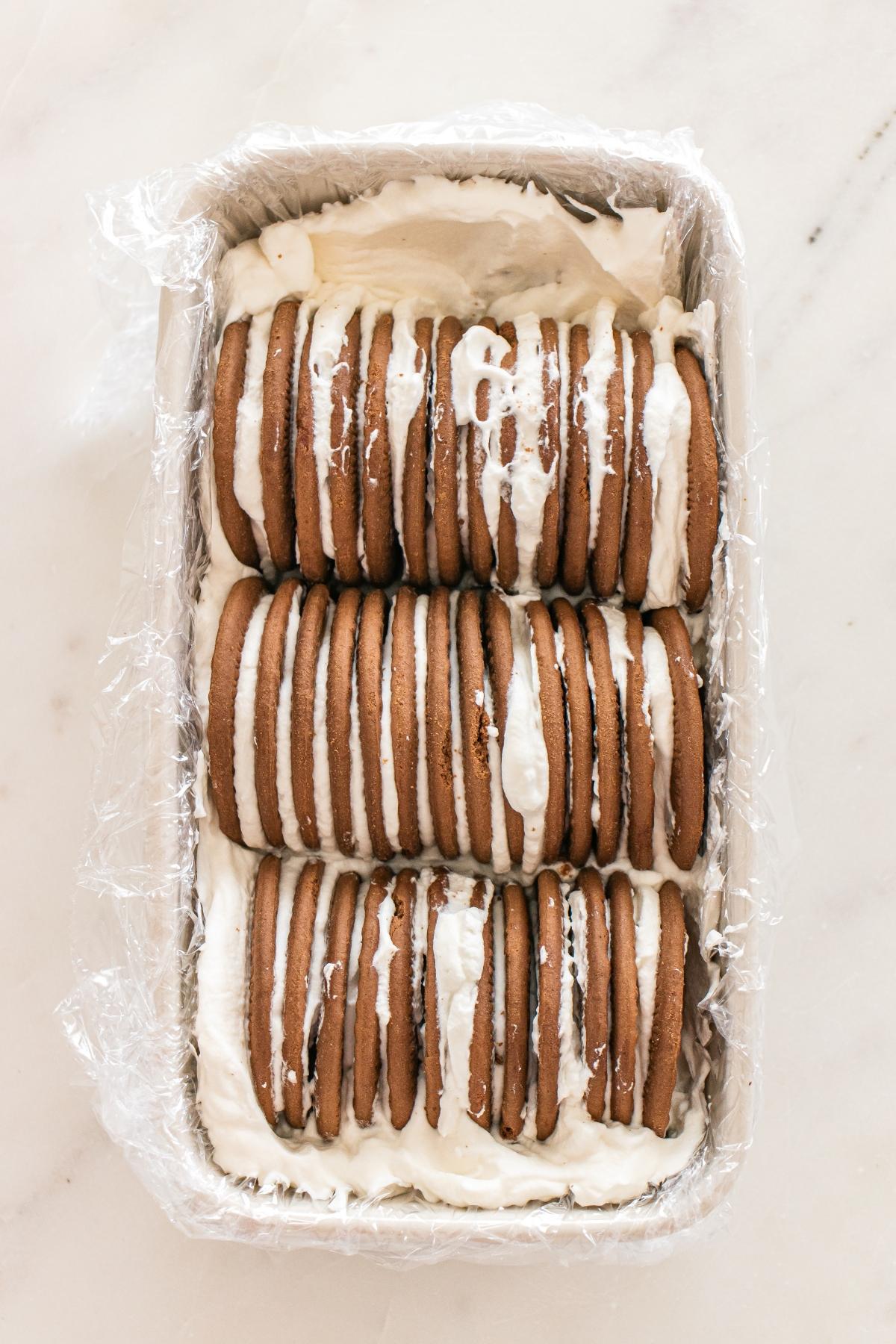 Chocolate wafer cookies layered with whipped cream in a loaf pan, lined with plastic wrap, on a white surface.