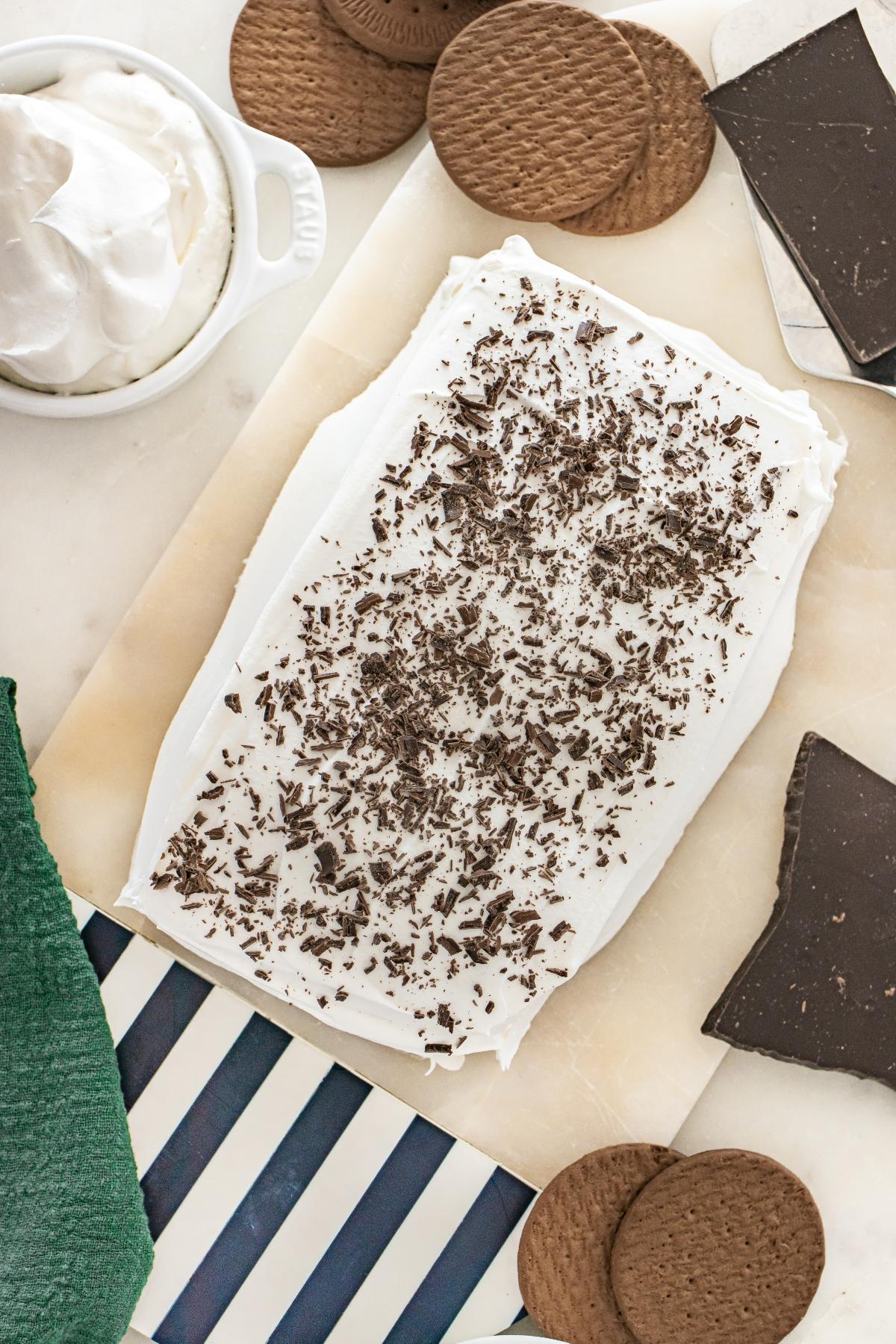 A rectangular icebox cake with whipped topping and chocolate shavings, surrounded by cookies and chocolate pieces.