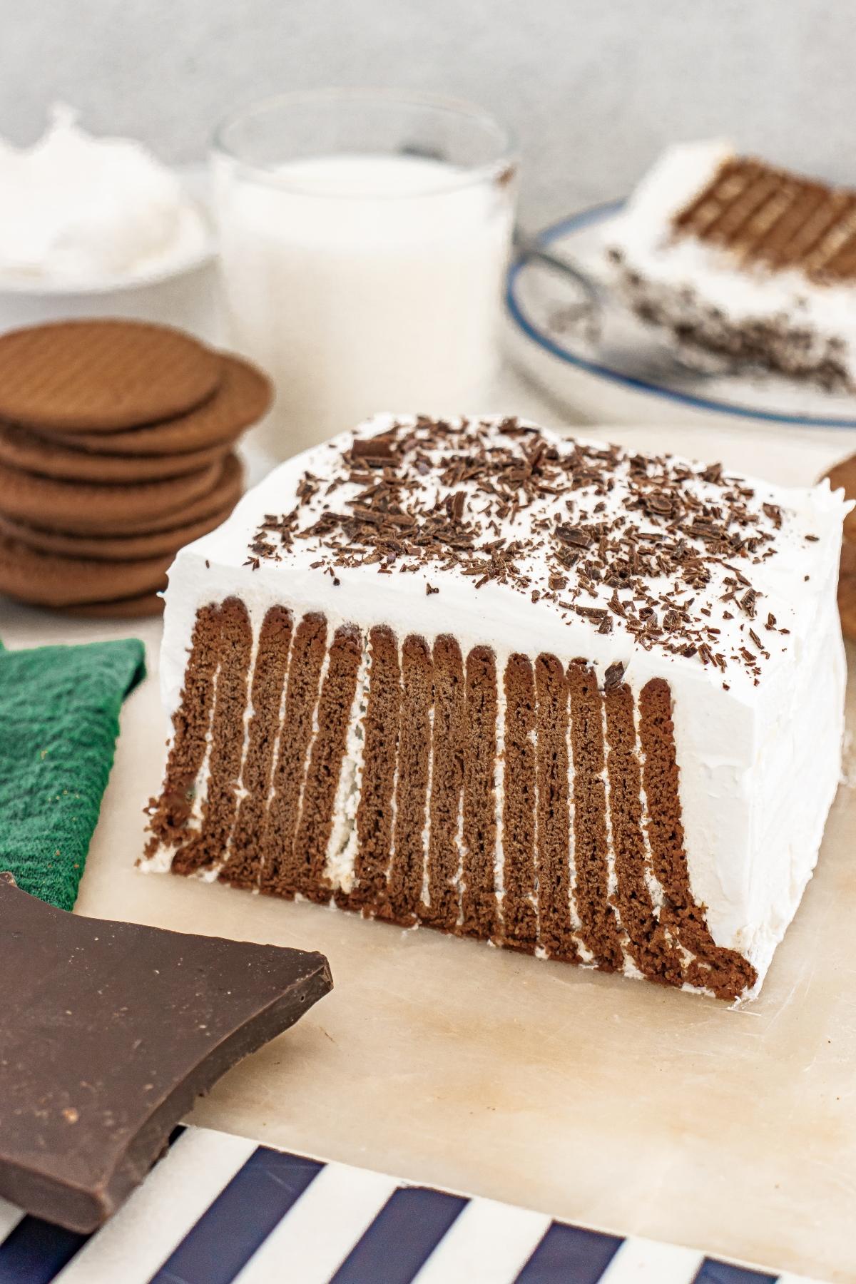 A slice of chocolate icebox cake with whipped cream and chocolate shavings on top, with cookies beside it.