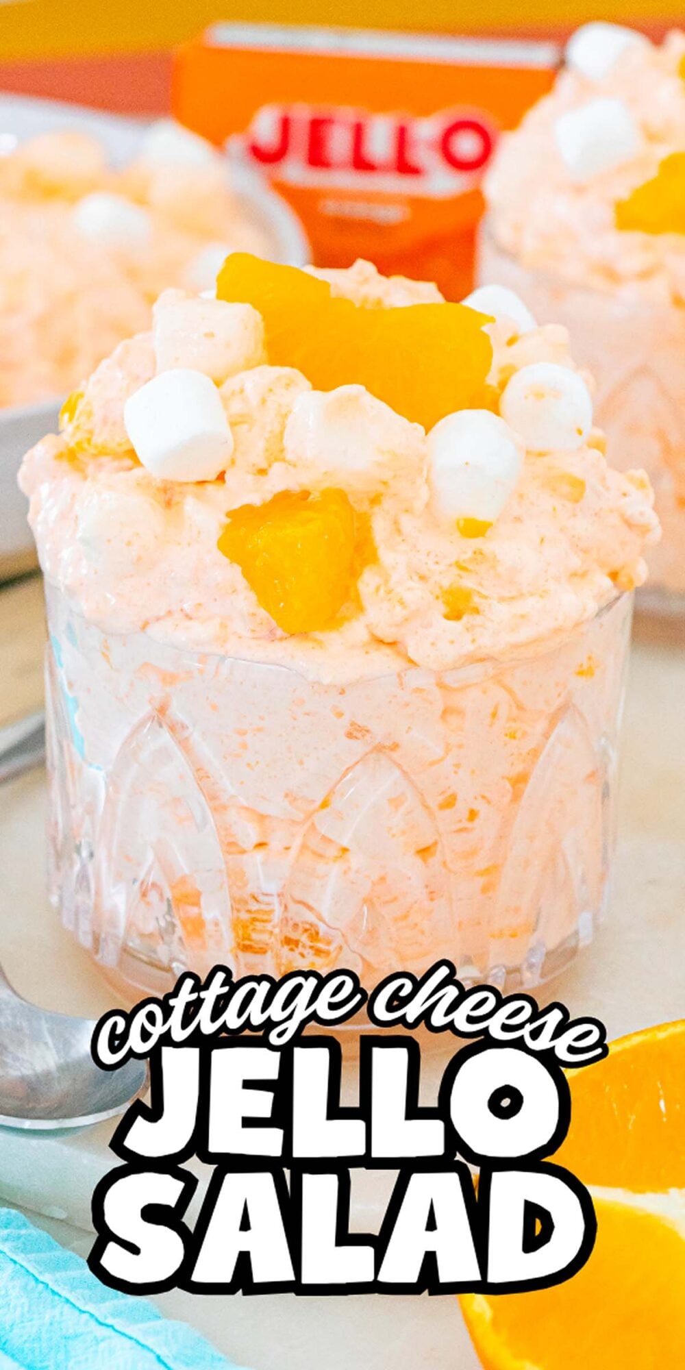 A glass of Cottage Cheese Jello Salad topped with orange slices and marshmallows, with an orange Jello box in the background.