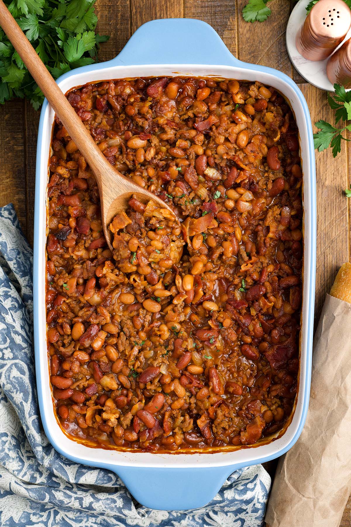 Cowboy baked beans in a blue casserole dish with a wooden spoon, garnished with herbs on a wooden table.