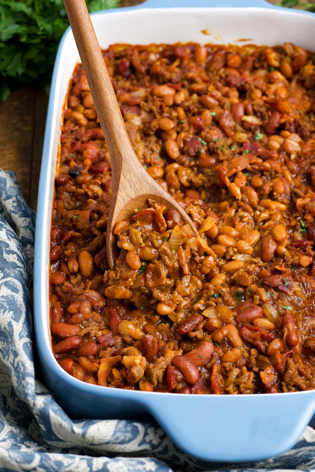 A wooden spoon scoops cowboy baked beans from a blue casserole dish filled with saucy, mixed beans.