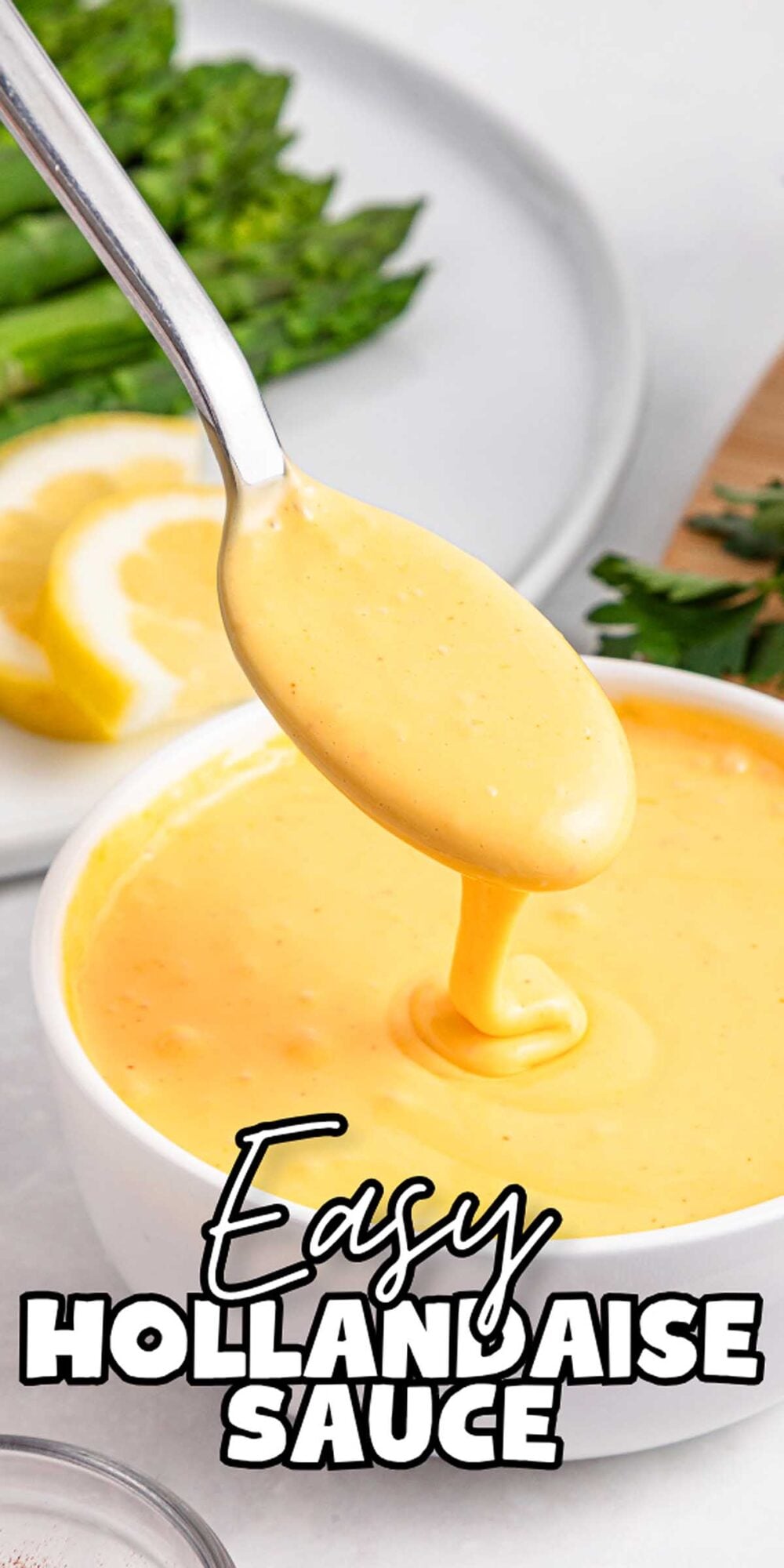 Spoon dipping into a bowl of creamy Easy Hollandaise Sauce, with lemon slices and greens in the background.