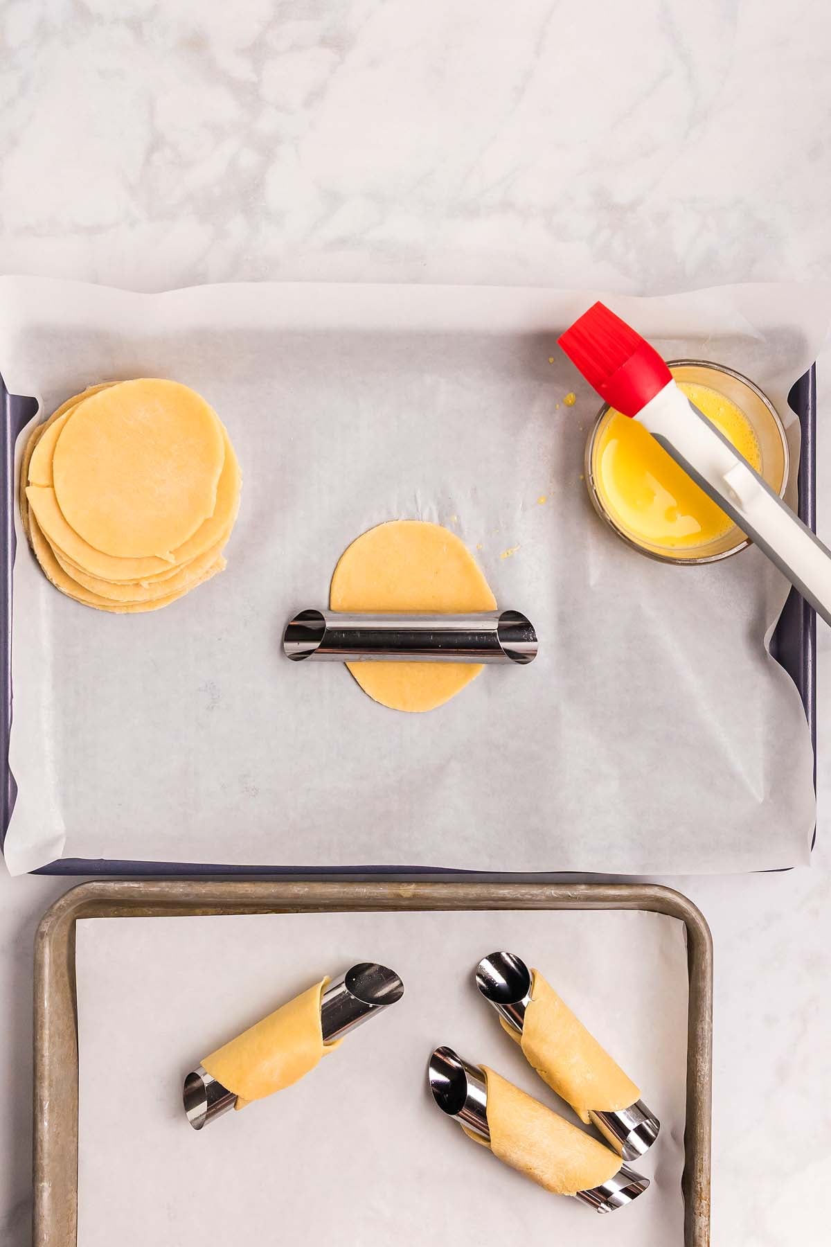 Round dough sheets, a pastry brush in egg wash, and homemade cannoli shells wrapped around metal tubes on a parchment-lined baking tray.