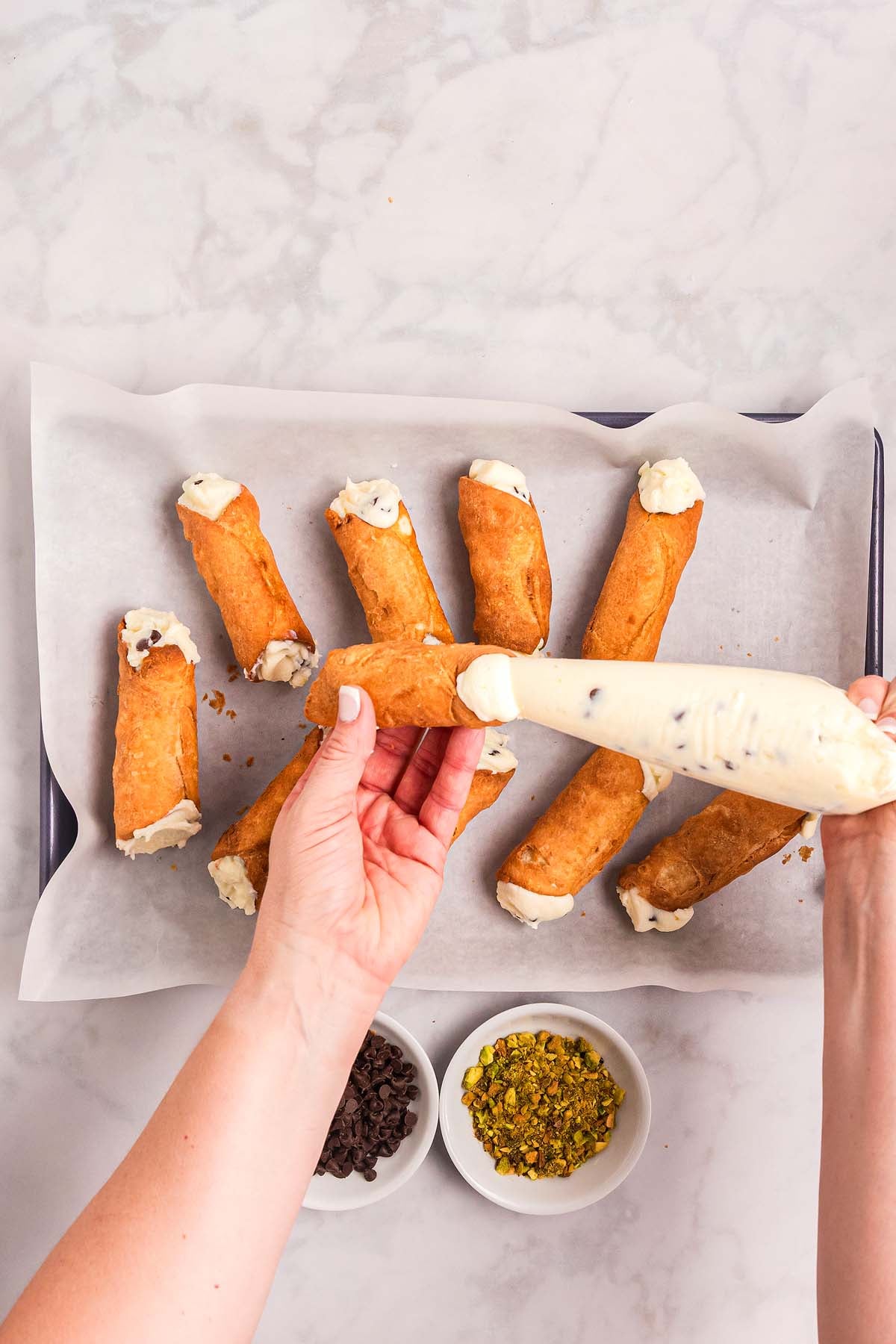 Hands filling a homemade cannoli shell with cream, surrounded by other delicious homemade cannoli on parchment paper.
