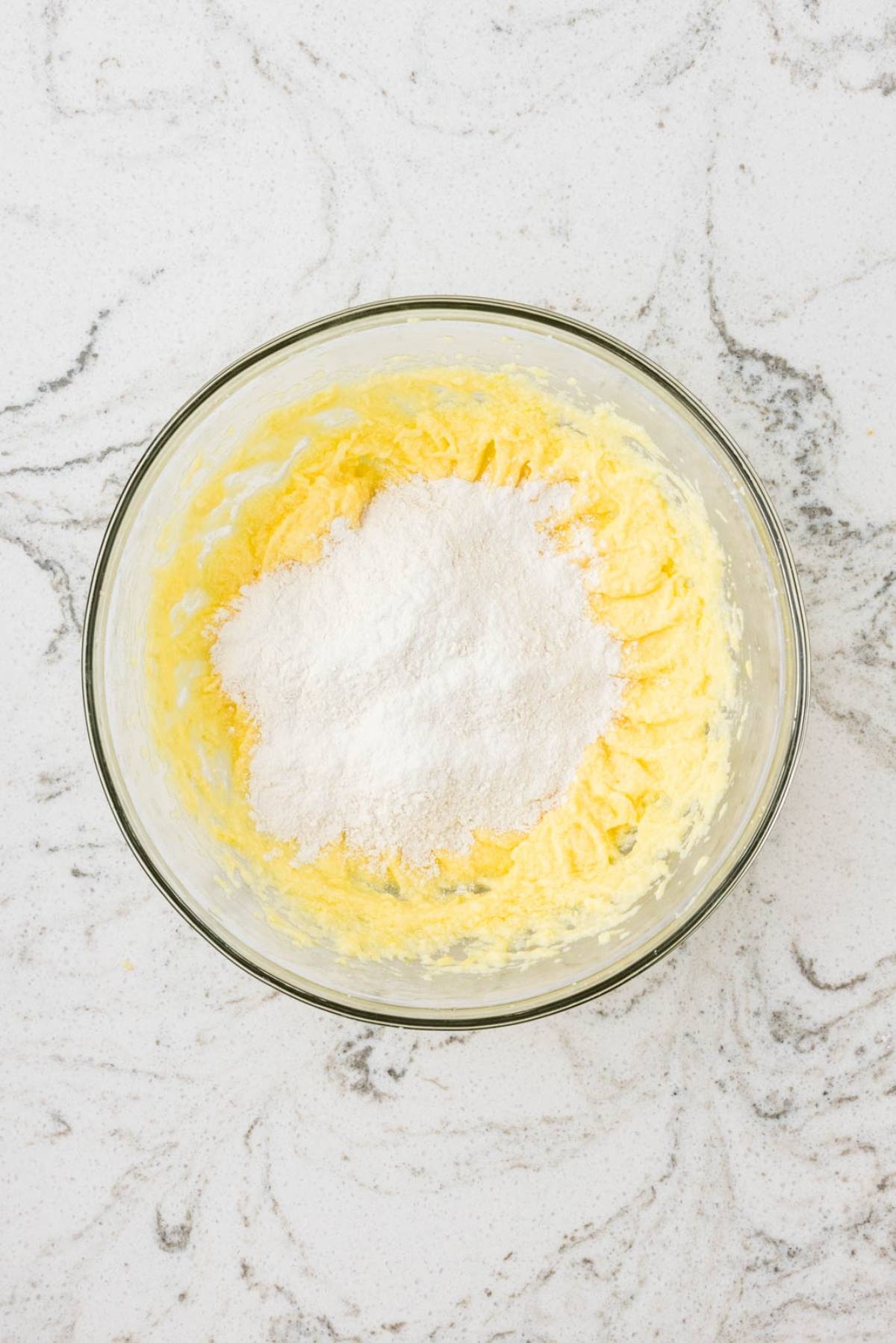 A glass bowl with creamed butter and sugar, topped with flour, on a white marble countertop.