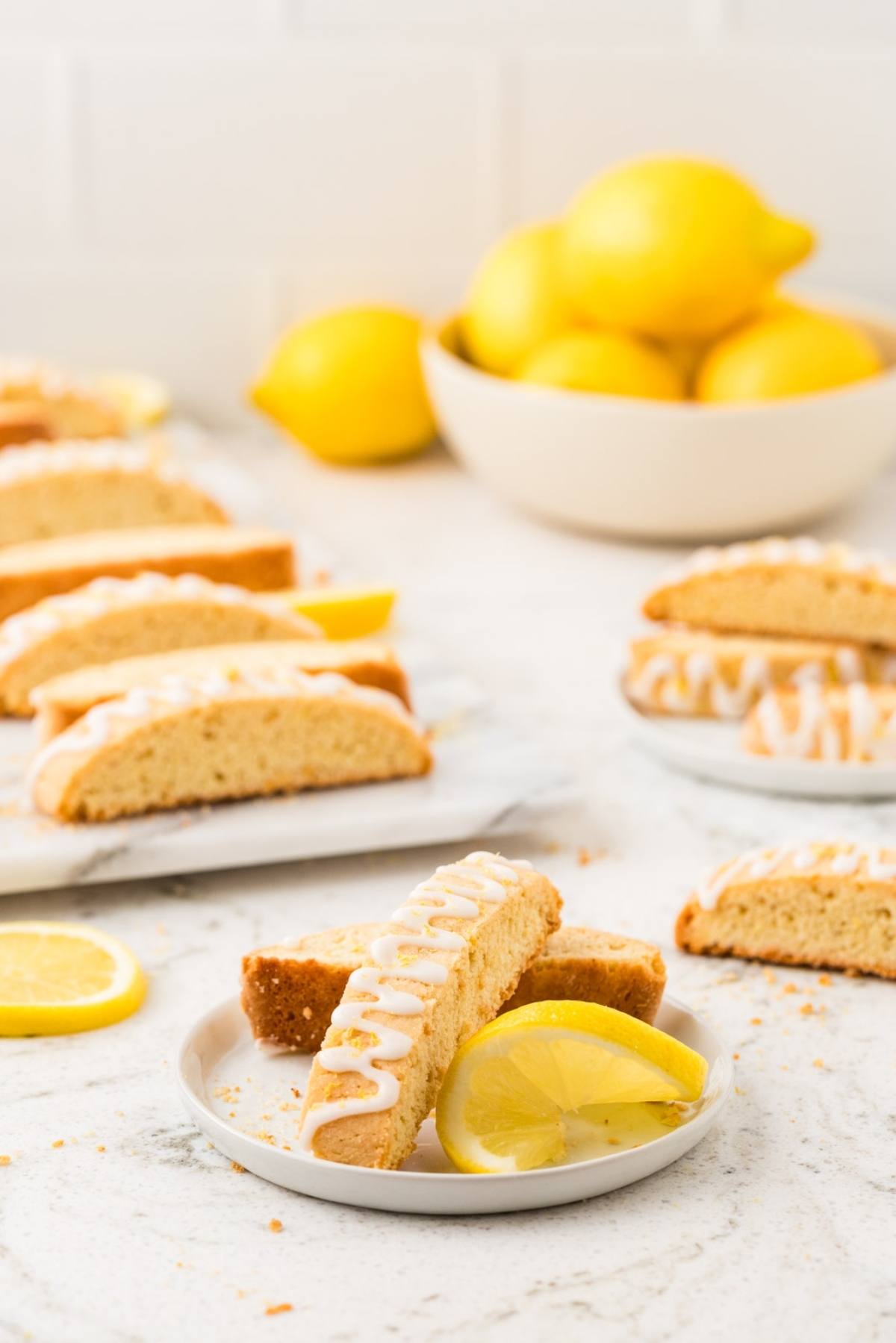 Lemon biscotti with icing on a plate, garnished with lemon slices; whole lemons in a bowl in the background.