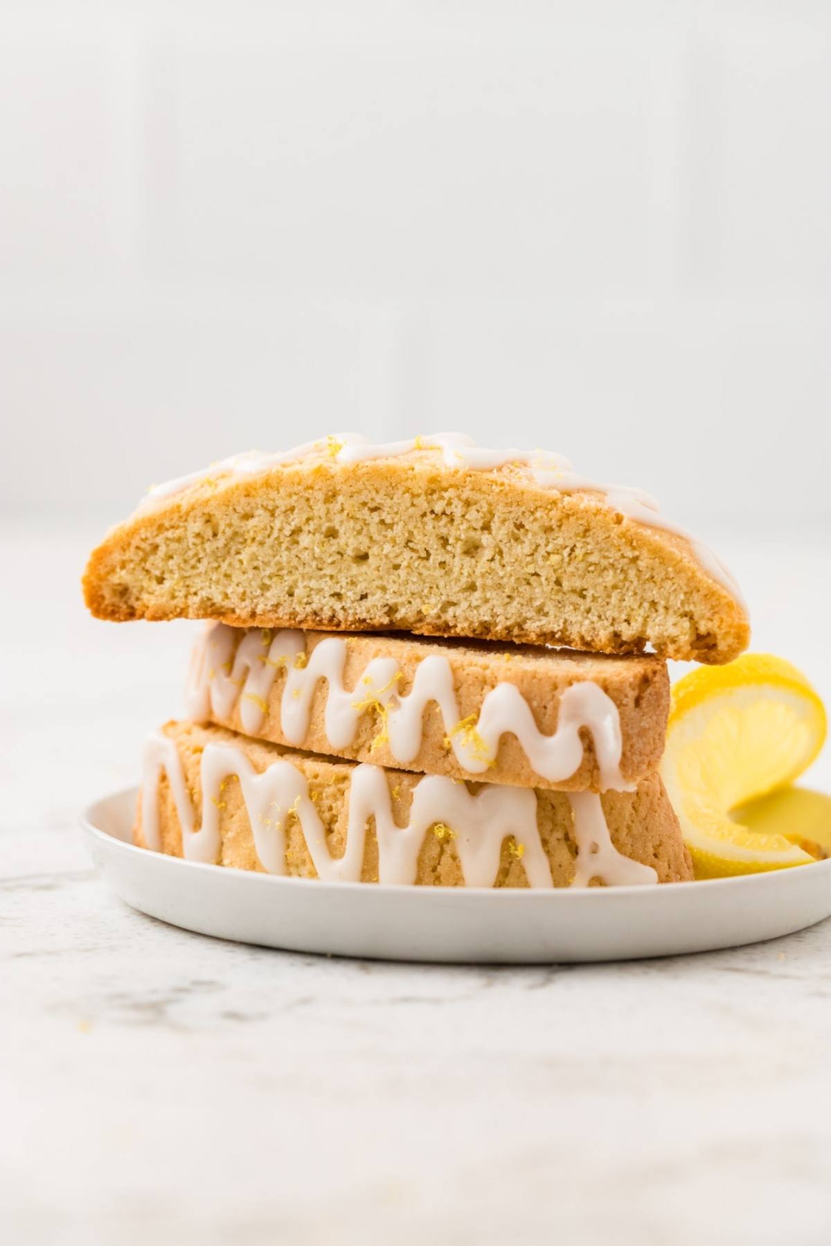 Three lemon biscotti with white icing are stacked on a plate with lemon slices beside them.