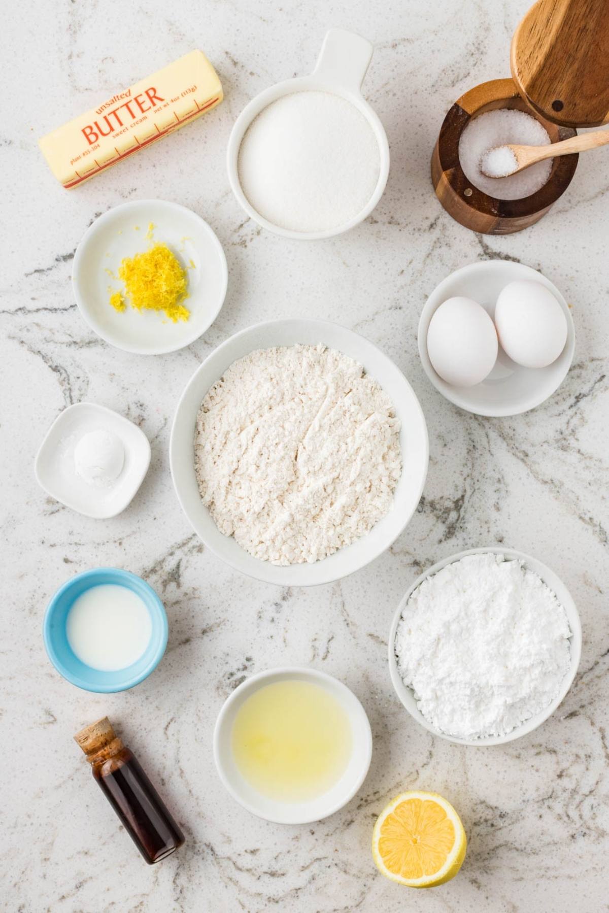 Baking ingredients on a marble counter: flour, sugar, butter, eggs, lemon, vanilla, milk, salt, and baking powder.