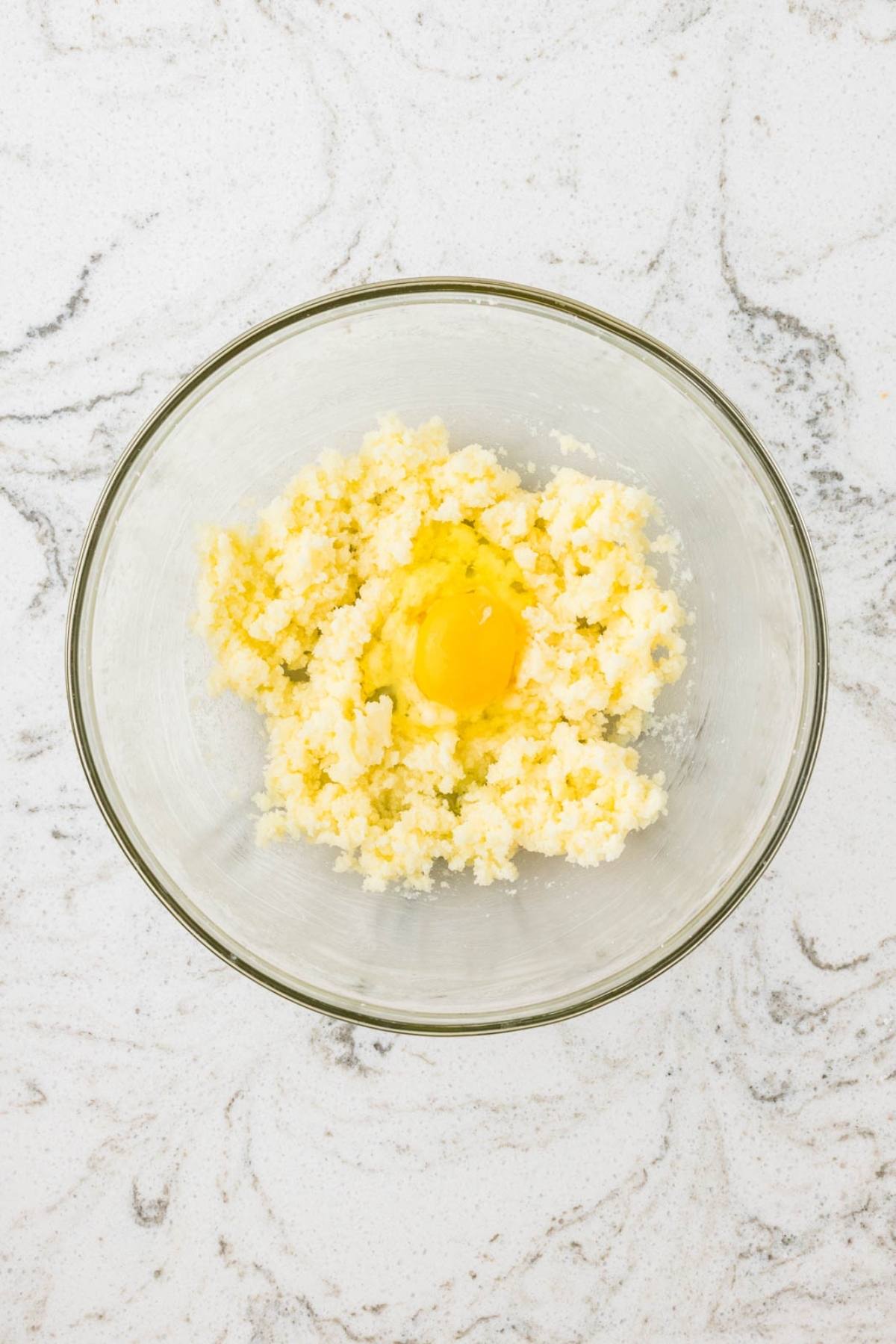 A glass bowl with creamed butter and sugar and a raw egg on a white marble surface.