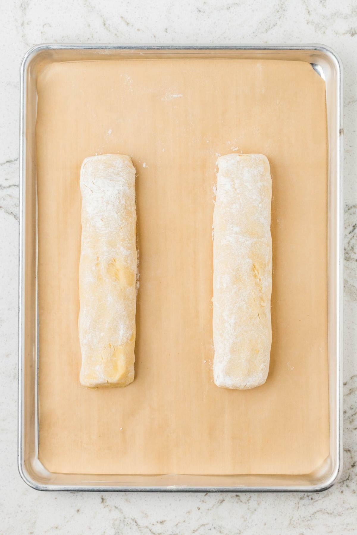 Two unbaked, floured dough logs on a parchment-lined baking sheet, on a light marble countertop.
