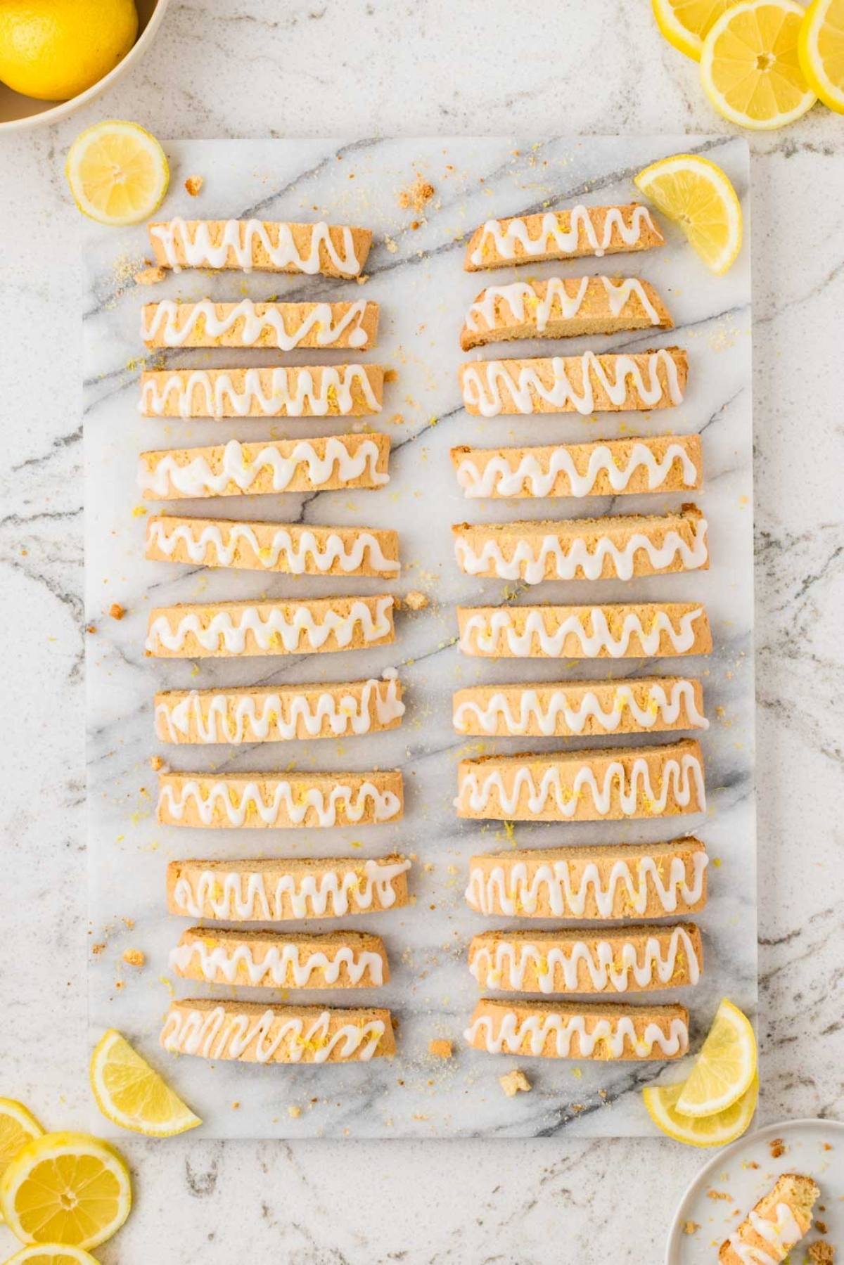 Rows of lemon biscotti with white icing drizzle on a marble board, surrounded by lemon slices.