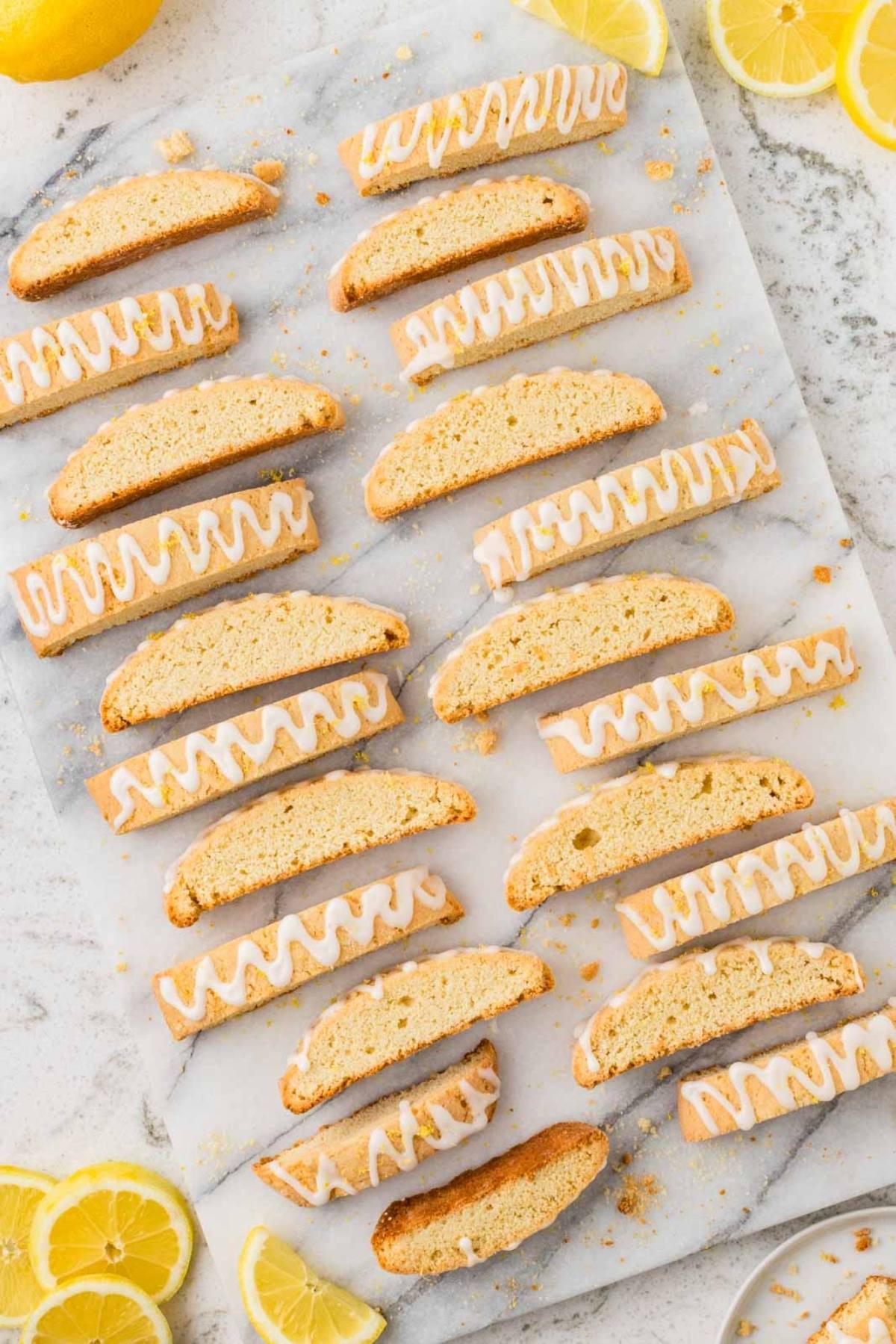 Rows of lemon biscotti with white icing drizzle on parchment, surrounded by lemon slices on a marble surface.
