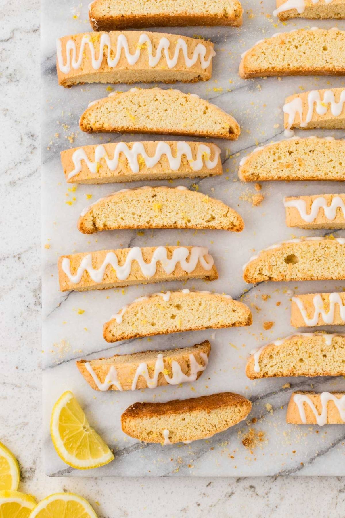Rows of lemon biscotti with white glaze on marble, with lemon slices beside them.