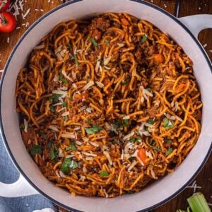 A one pot pasta of spaghetti with meat sauce, topped with grated cheese and parsley, sits on a wooden table surrounded by fresh ingredients.