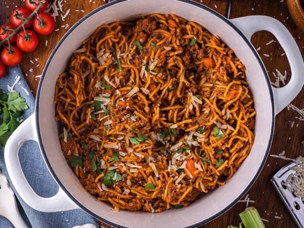 A one pot pasta of spaghetti with meat sauce, topped with grated cheese and parsley, sits on a wooden table surrounded by fresh ingredients.