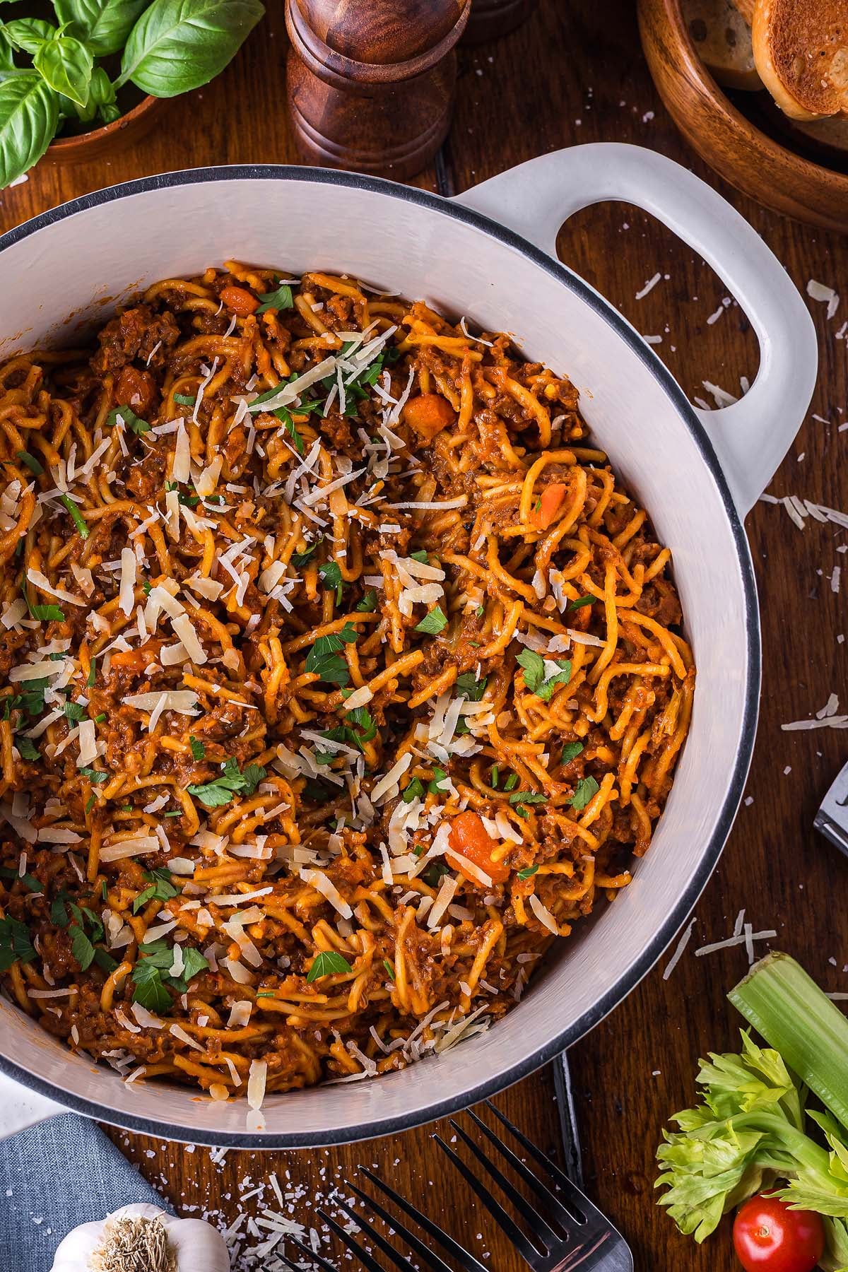 A pot of one pot pasta bolognese topped with grated cheese and parsley sits on a wooden table surrounded by fresh ingredients.