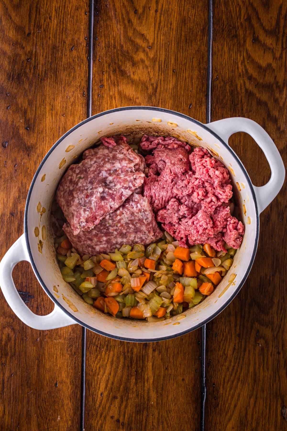 White pot with chopped carrots, celery, onion, and two portions of raw ground meat on a wooden table.
