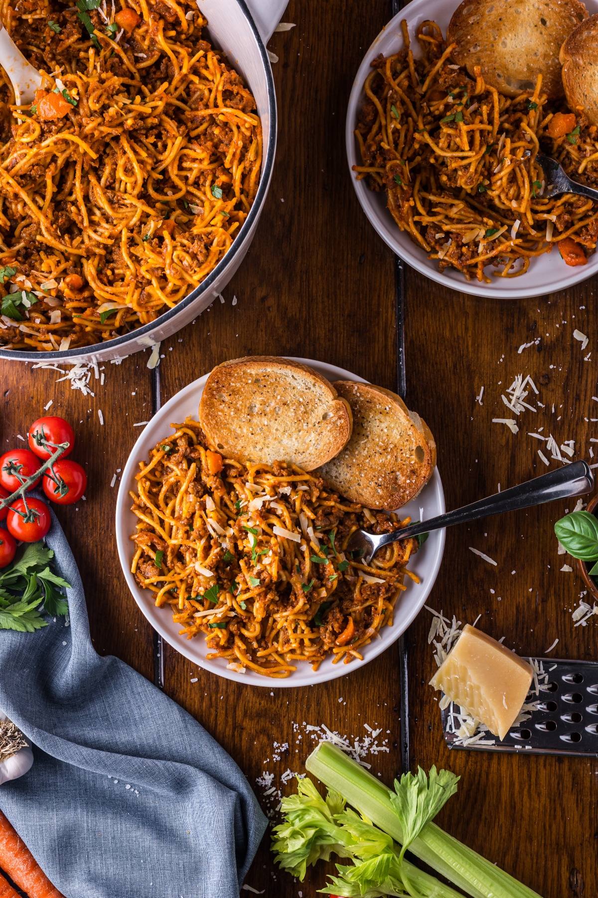 Plates of One pot pasta spaghetti with tomato sauce and toast on a wooden table, surrounded by cheese, tomatoes, and celery.