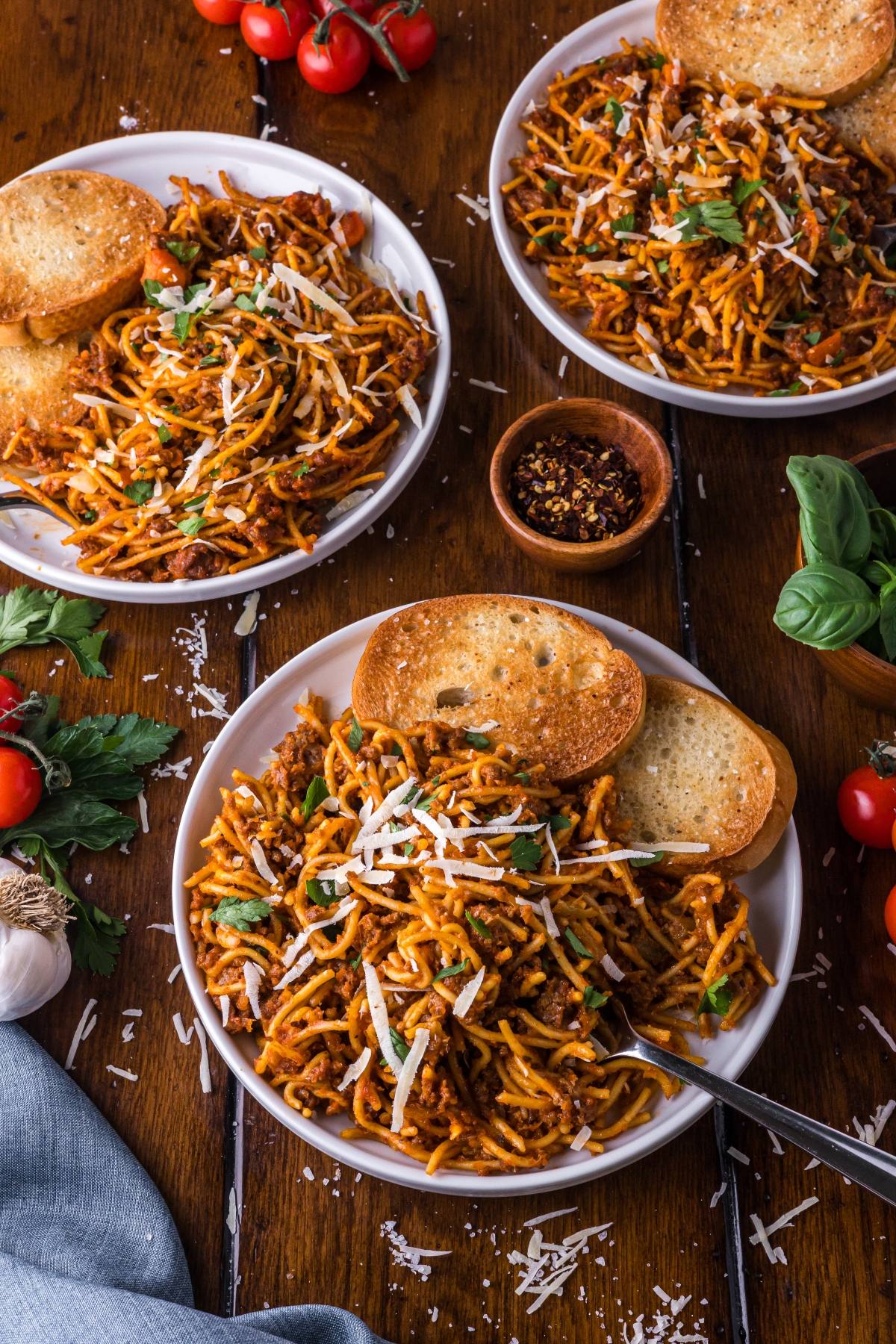 Three plates of One pot pasta spaghetti with tomato sauce, parmesan, and garlic bread on a wooden table, garnished with basil.