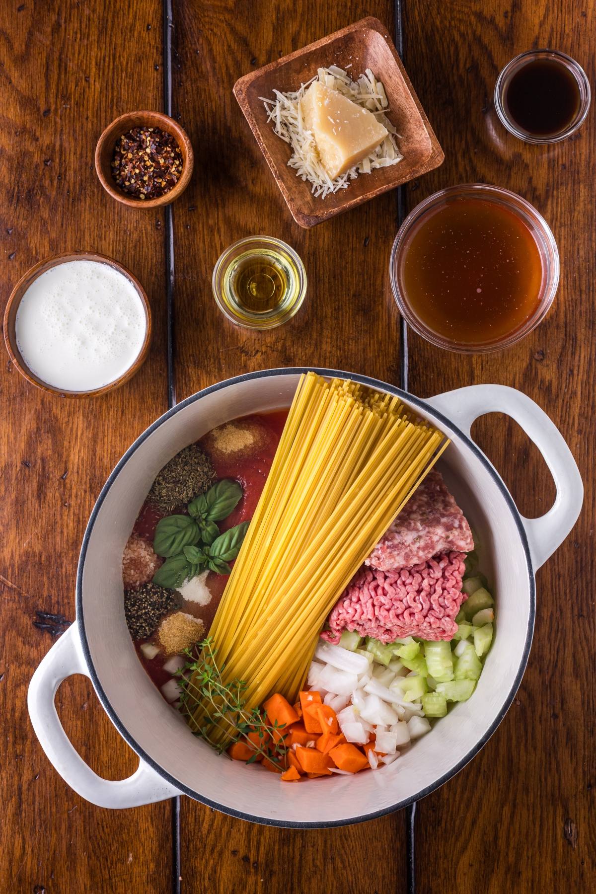 Uncooked pasta, meats, and vegetables in a pot, surrounded by spices, cheese, and liquids on a wooden table.