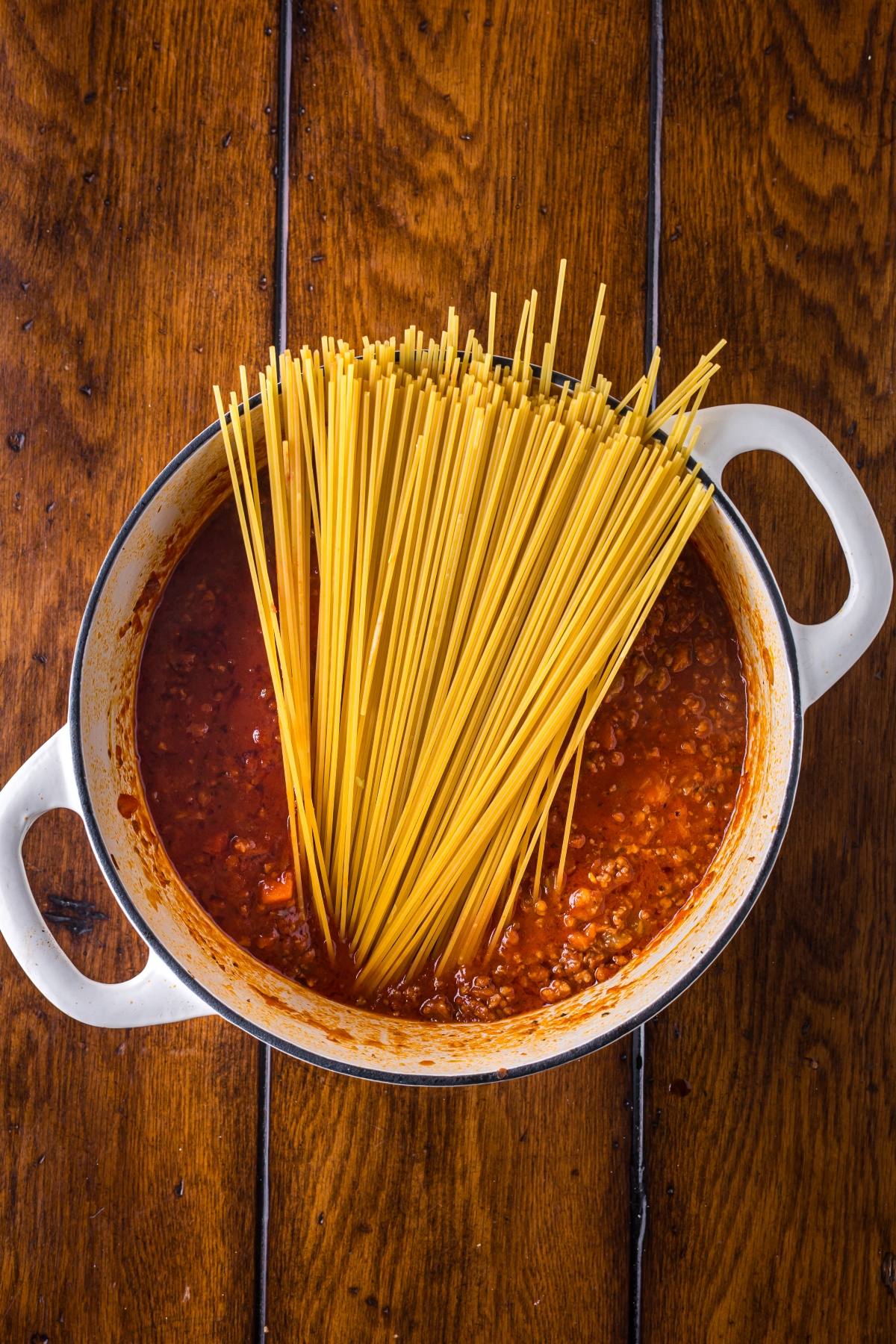 Uncooked spaghetti is placed in a pot of red meat sauce on a wooden table.