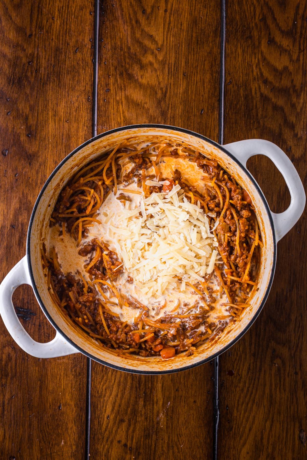 A pot of creamy spaghetti with meat sauce, topped with shredded cheese, on a wooden table.