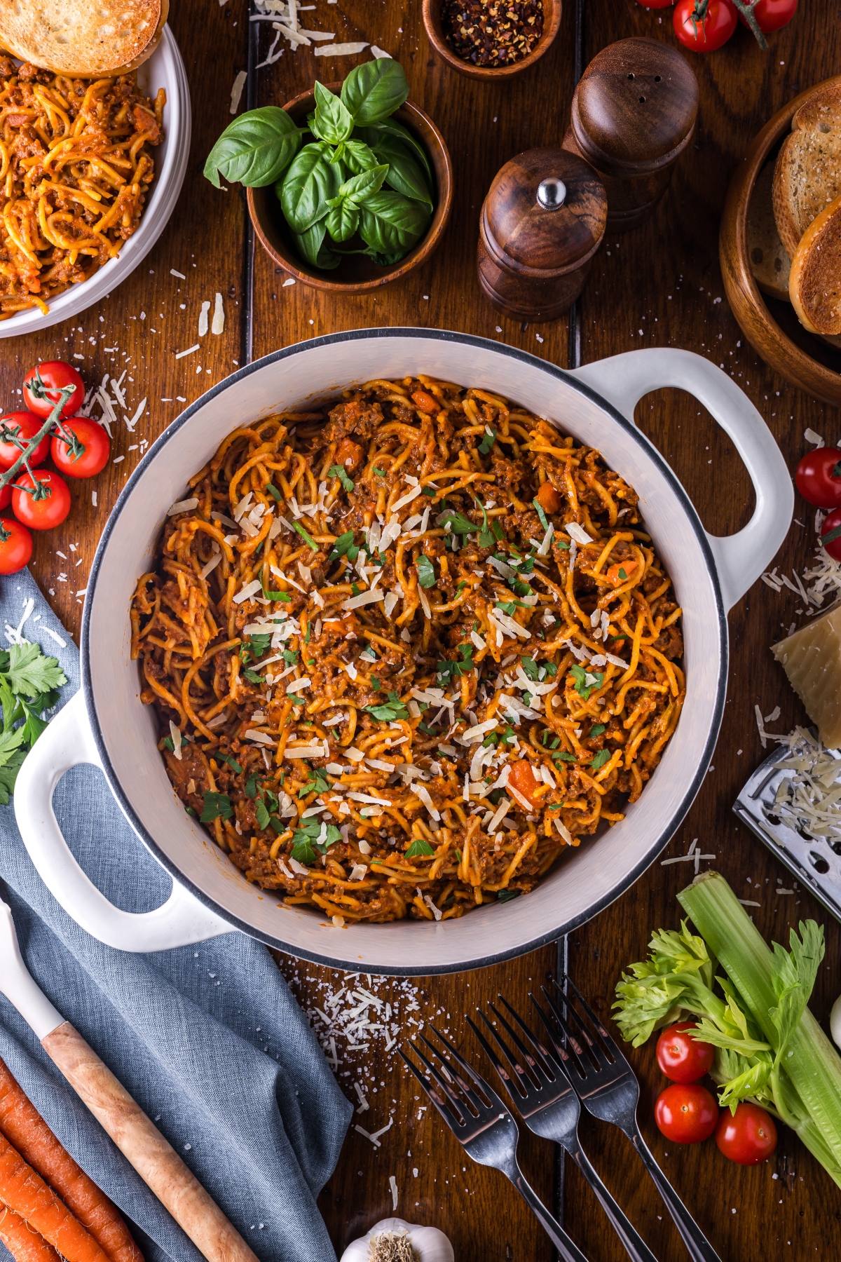A One pot pasta of spaghetti with meat sauce, topped with grated cheese and herbs, surrounded by fresh ingredients and utensils.