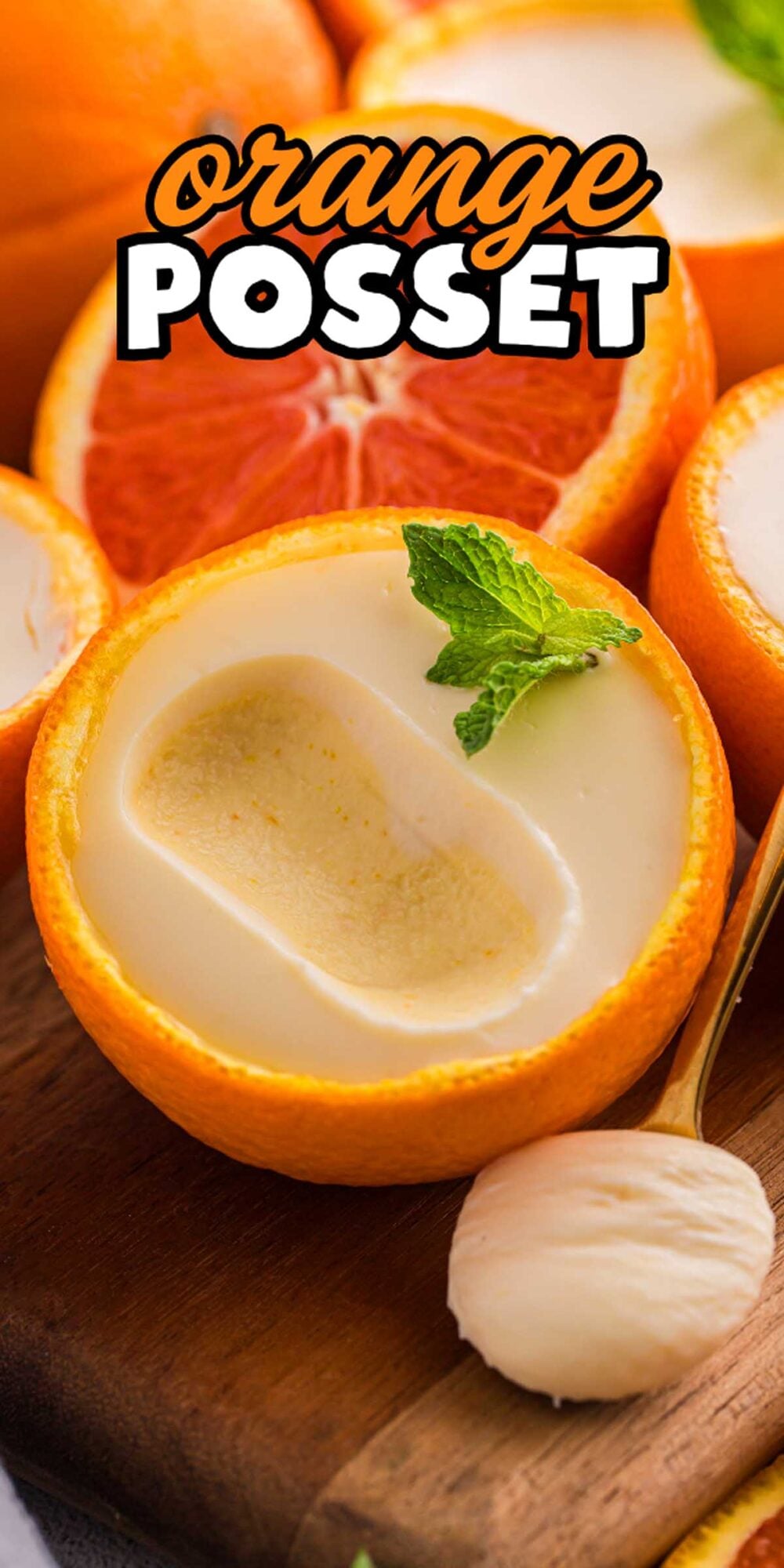 Orange posset served in a hollowed orange, topped with mint, with a spoonful of orange posset beside it on a wooden board.