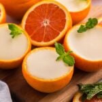 Halved oranges filled with creamy orange posset, garnished with mint leaves, rest elegantly on a wooden board.