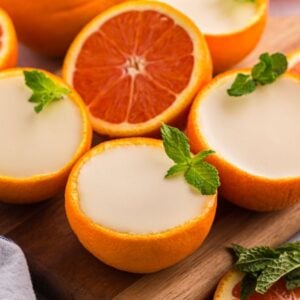 Halved oranges filled with creamy orange posset, garnished with mint leaves, rest elegantly on a wooden board.