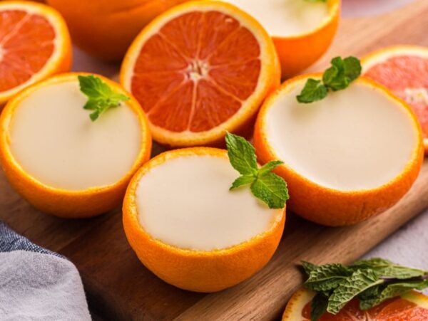 Halved oranges filled with creamy orange posset, garnished with mint leaves, rest elegantly on a wooden board.
