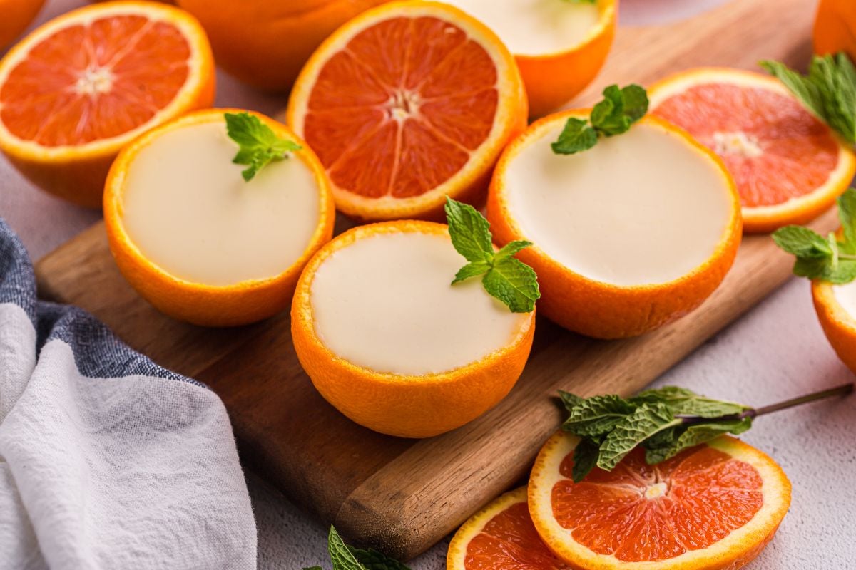 Halved oranges filled with creamy orange posset, garnished with mint, on a wooden board with fresh orange slices.