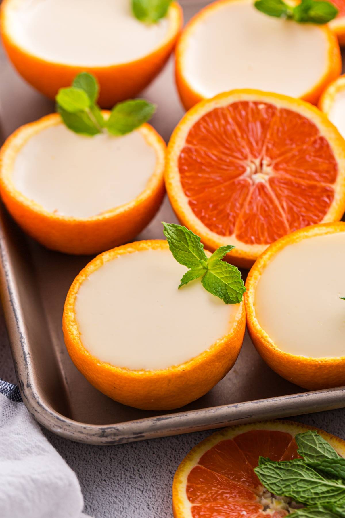 Orange posset halves filled with creamy dessert, garnished with mint leaves, on a tray beside orange slices.