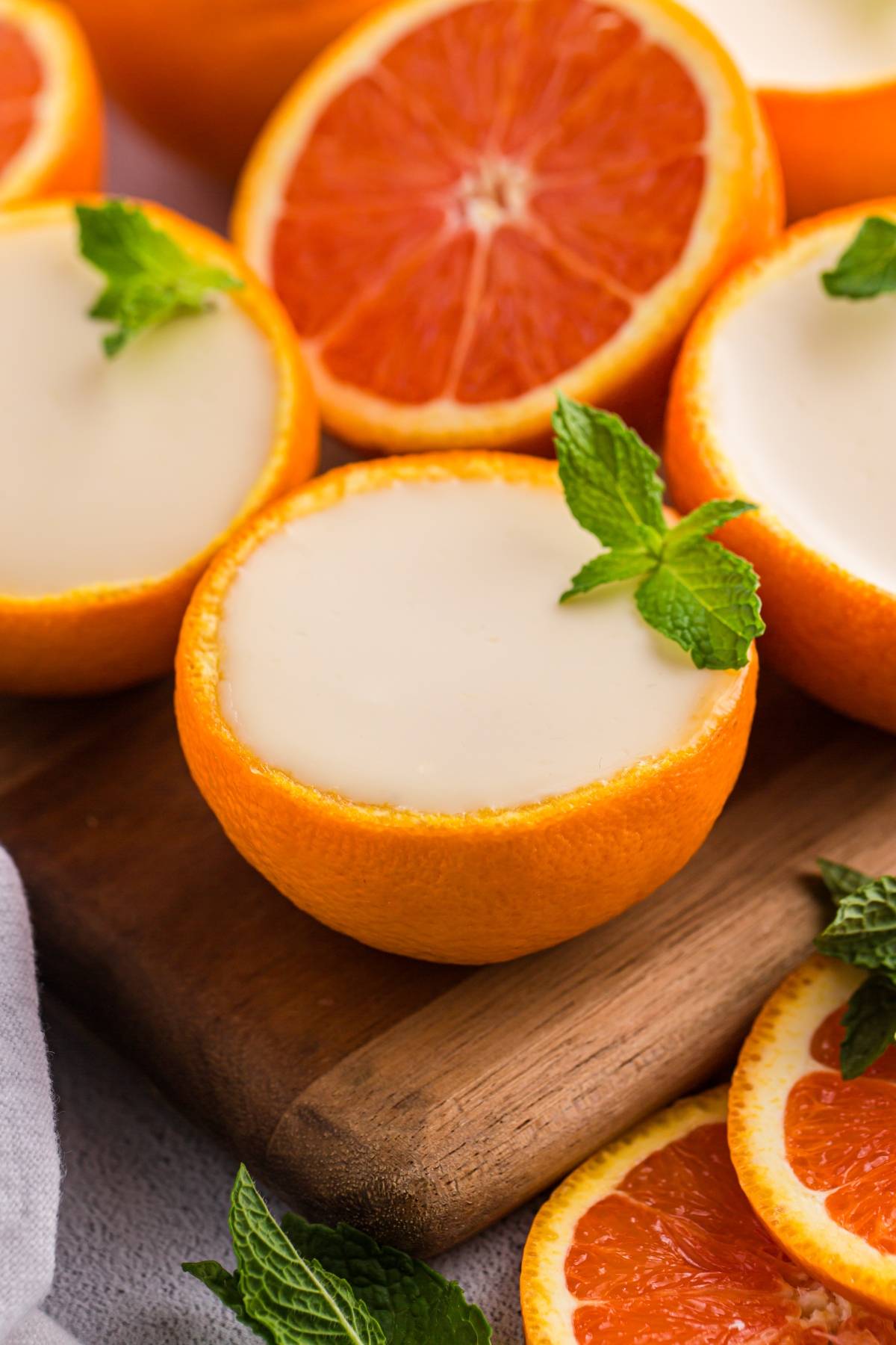 Orange posset halves filled with creamy dessert, garnished with fresh mint, on a wooden board with orange slices.