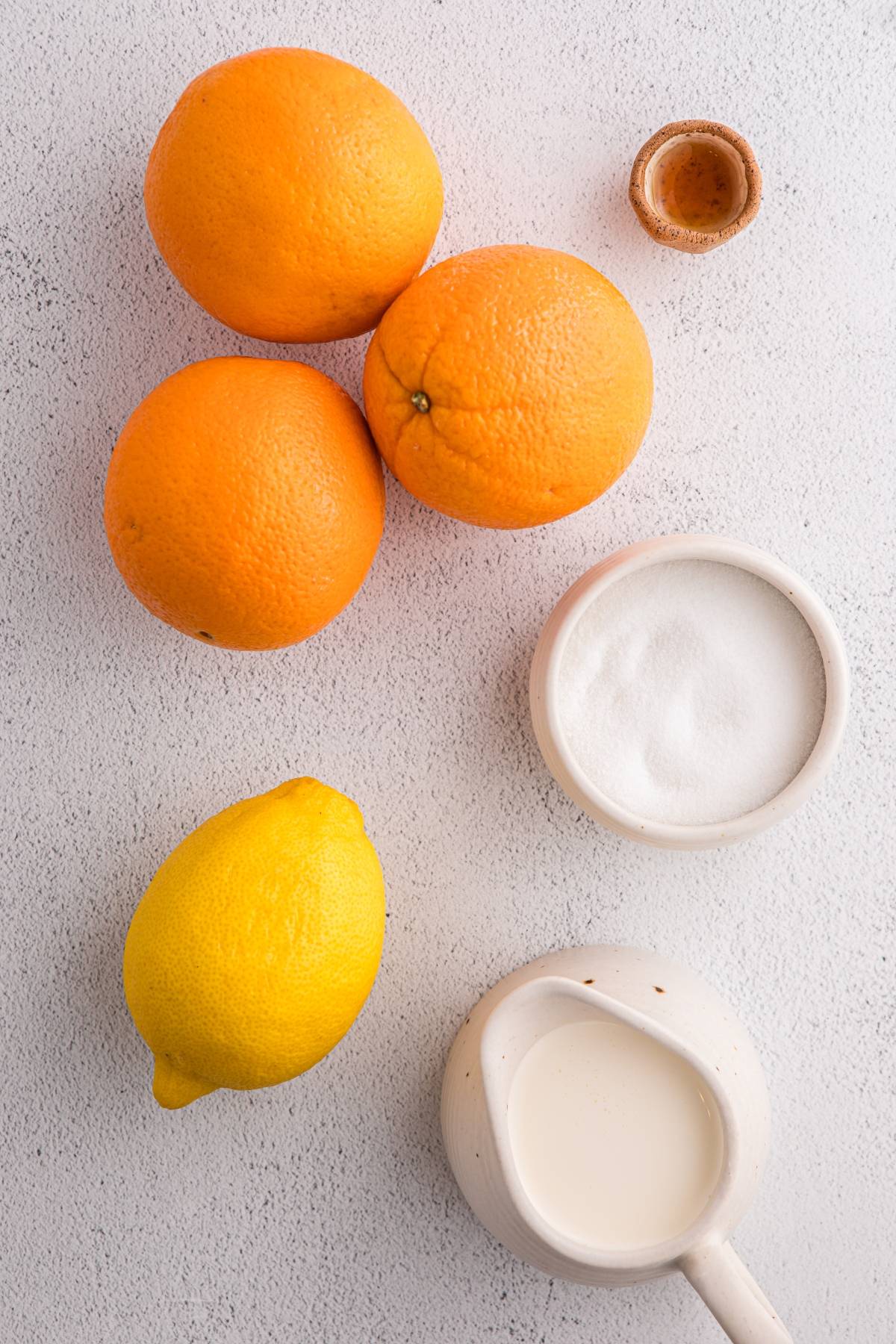 Three oranges, a lemon, a small bowl of sugar, a small pitcher of cream, and a small cup of vanilla on a white surface.