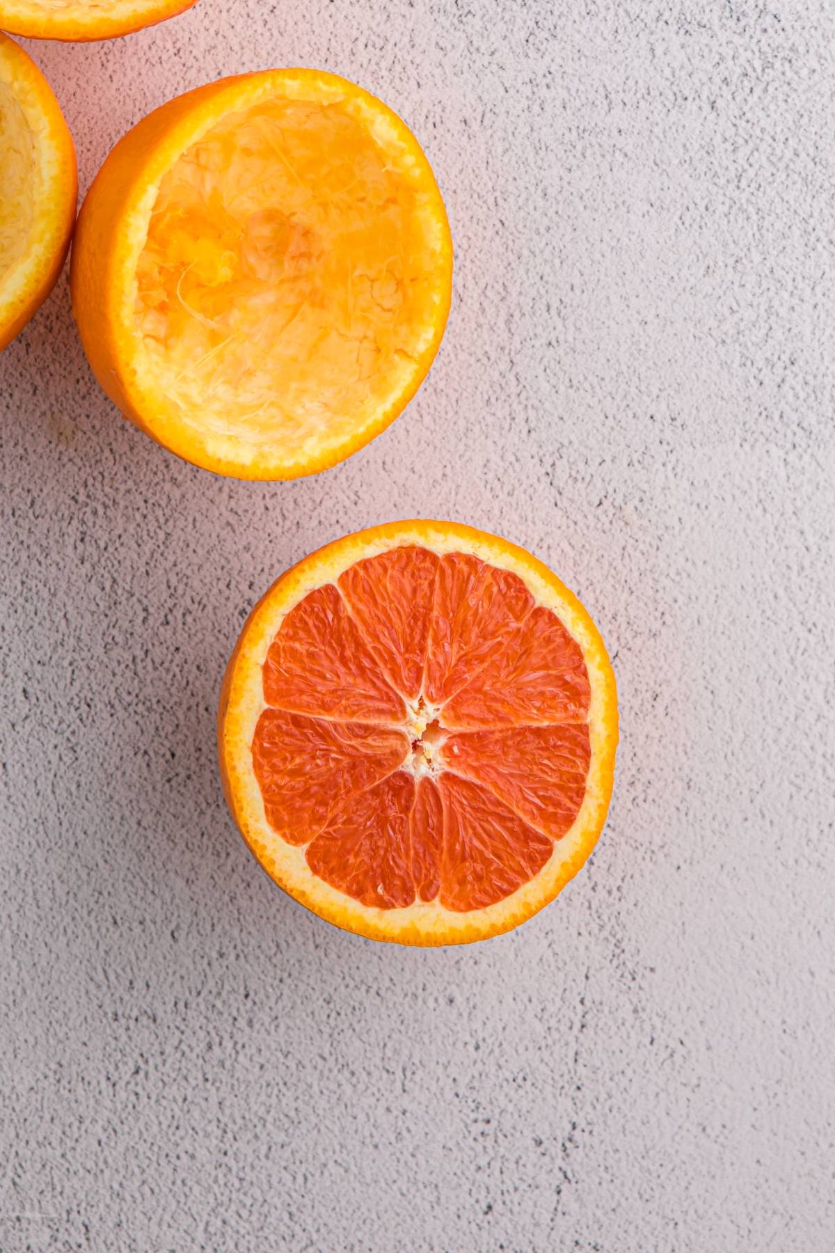 Half of a juicy orange with a vibrant interior, next to two squeezed orange halves on a light surface.