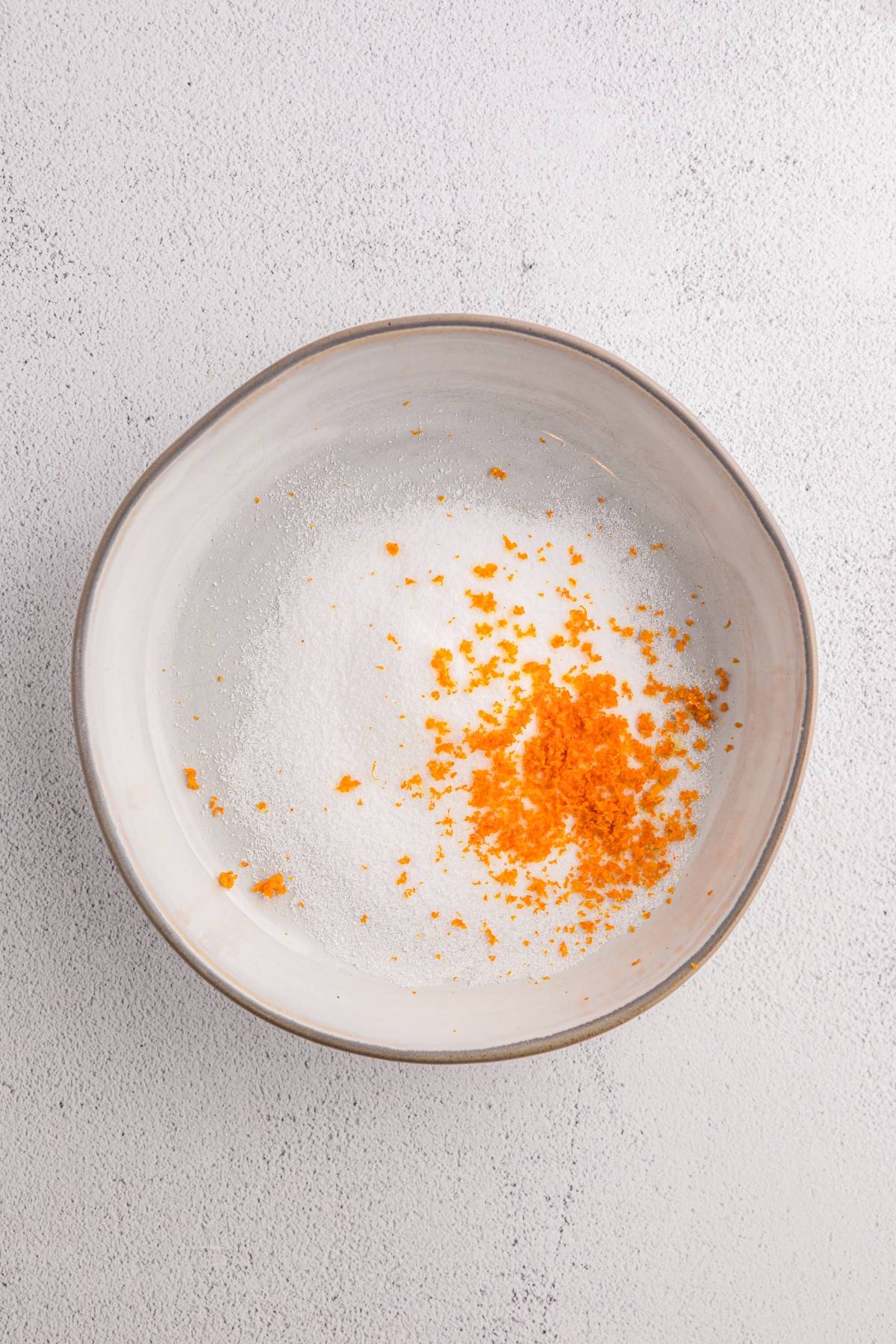 A bowl with white sugar and a small pile of orange zest on a light textured surface.