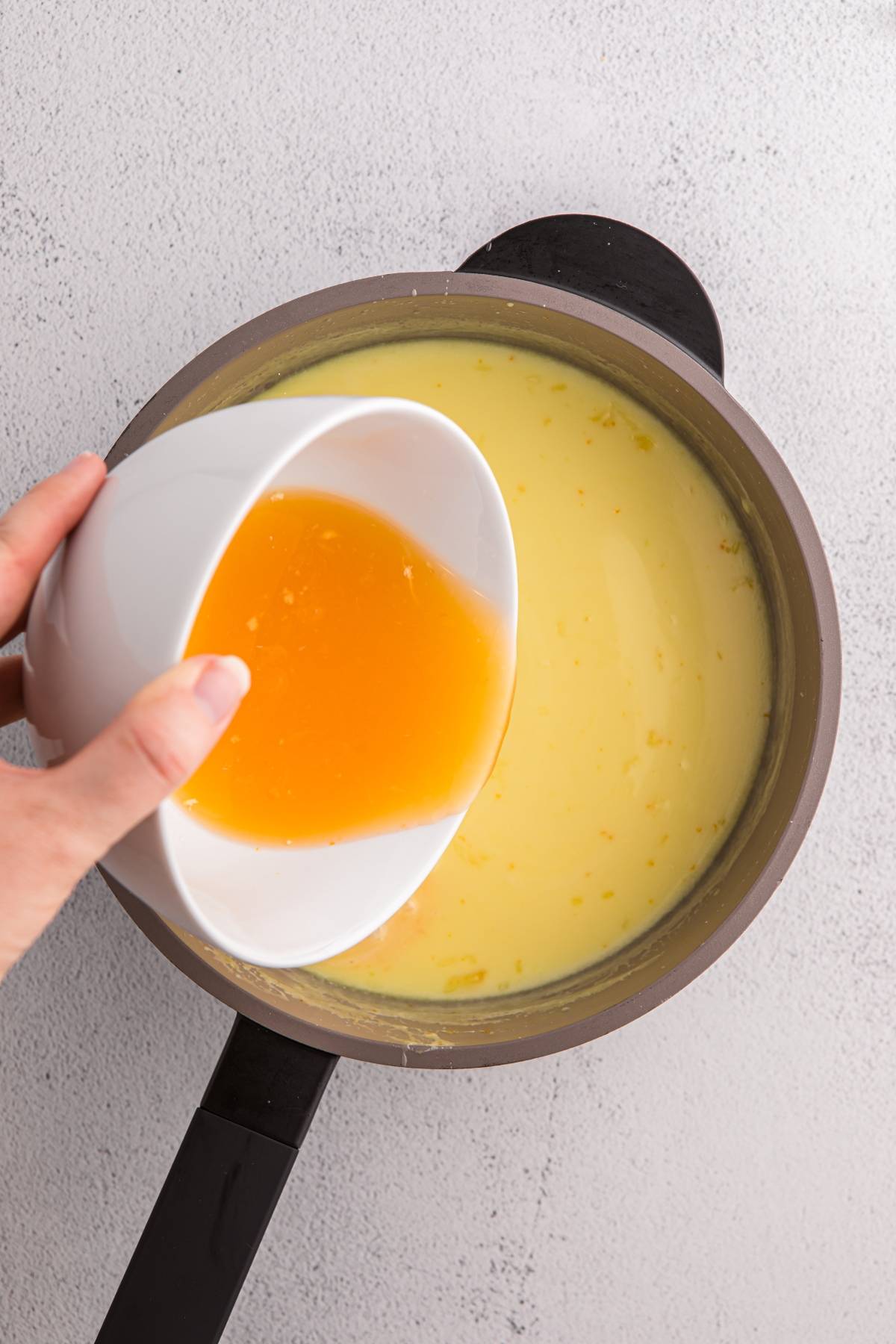 A hand pours orange liquid from a bowl into a pot of creamy yellow mixture on a light surface.