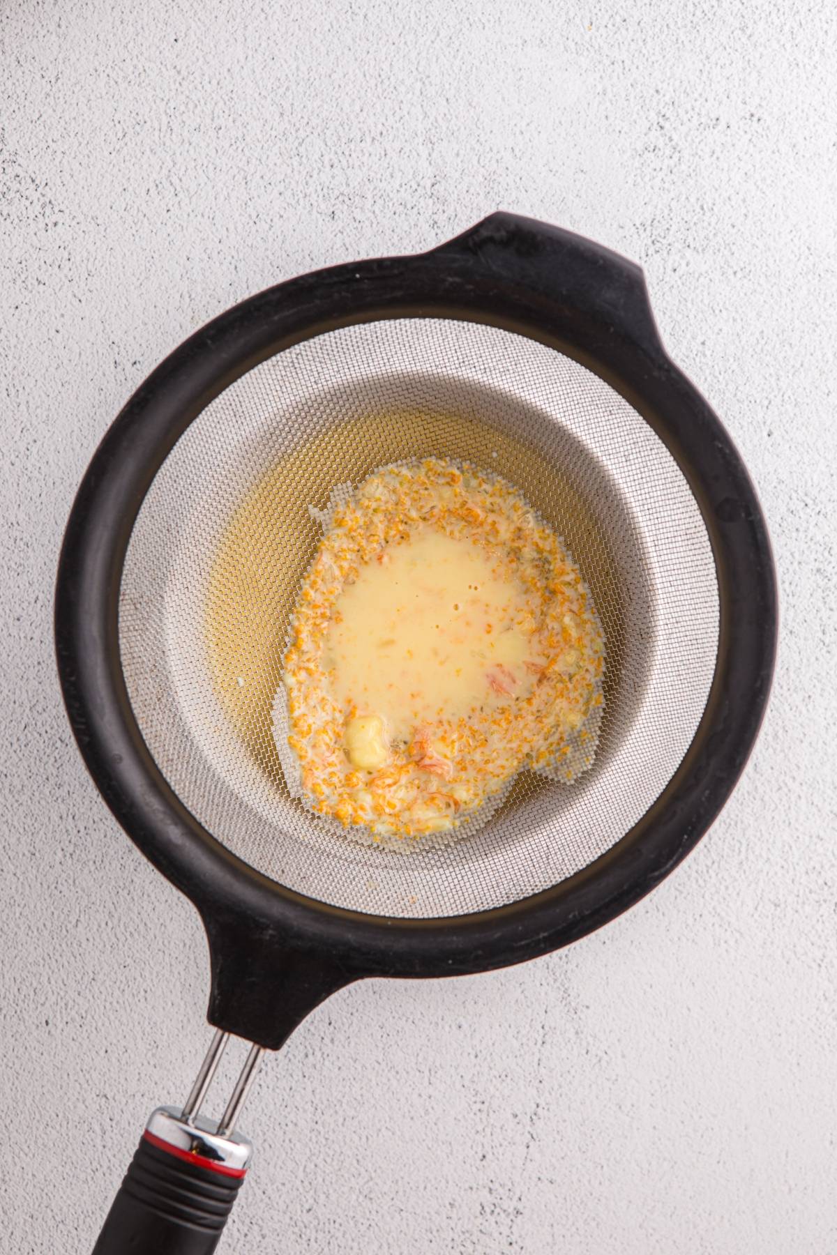 A metal strainer with a black handle holds a mixture of liquid and orange zest on a light surface.