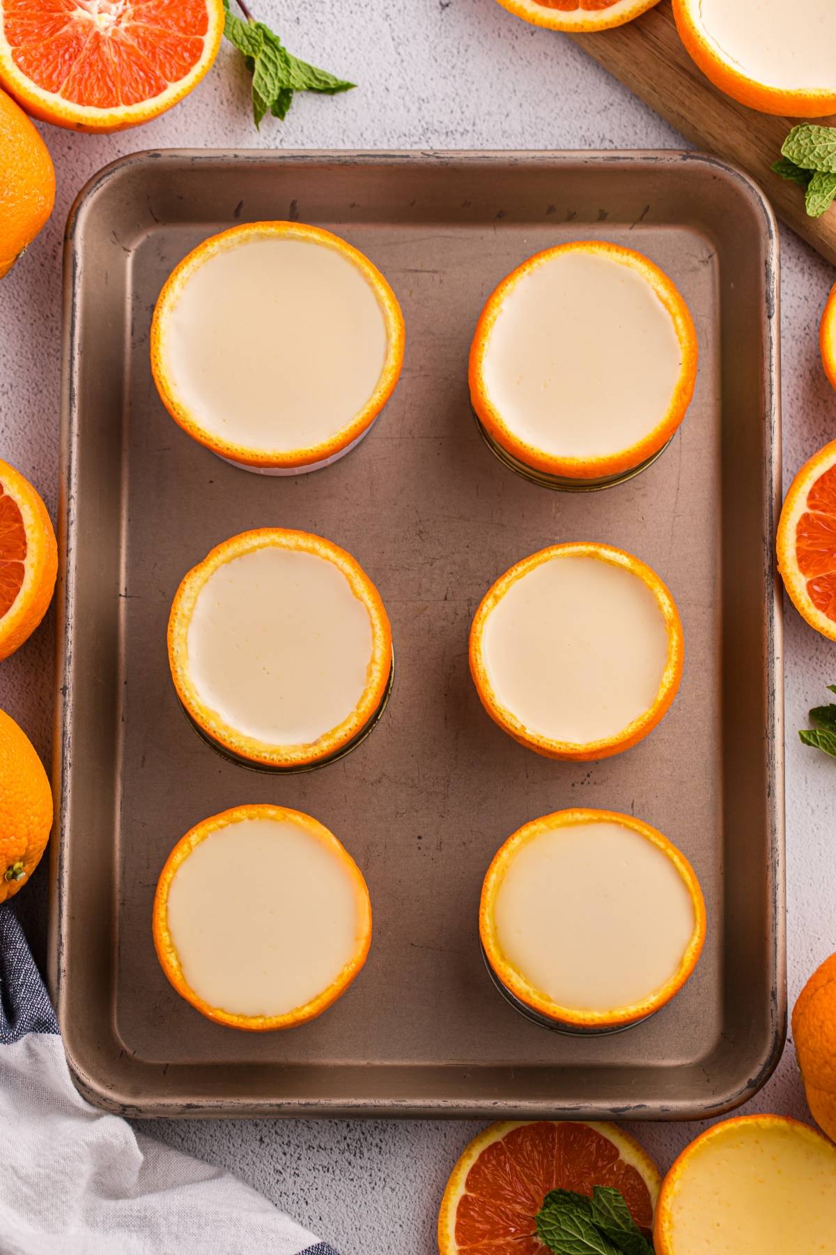 Six orange halves filled with creamy dessert on a baking tray, surrounded by fresh orange slices and mint leaves.