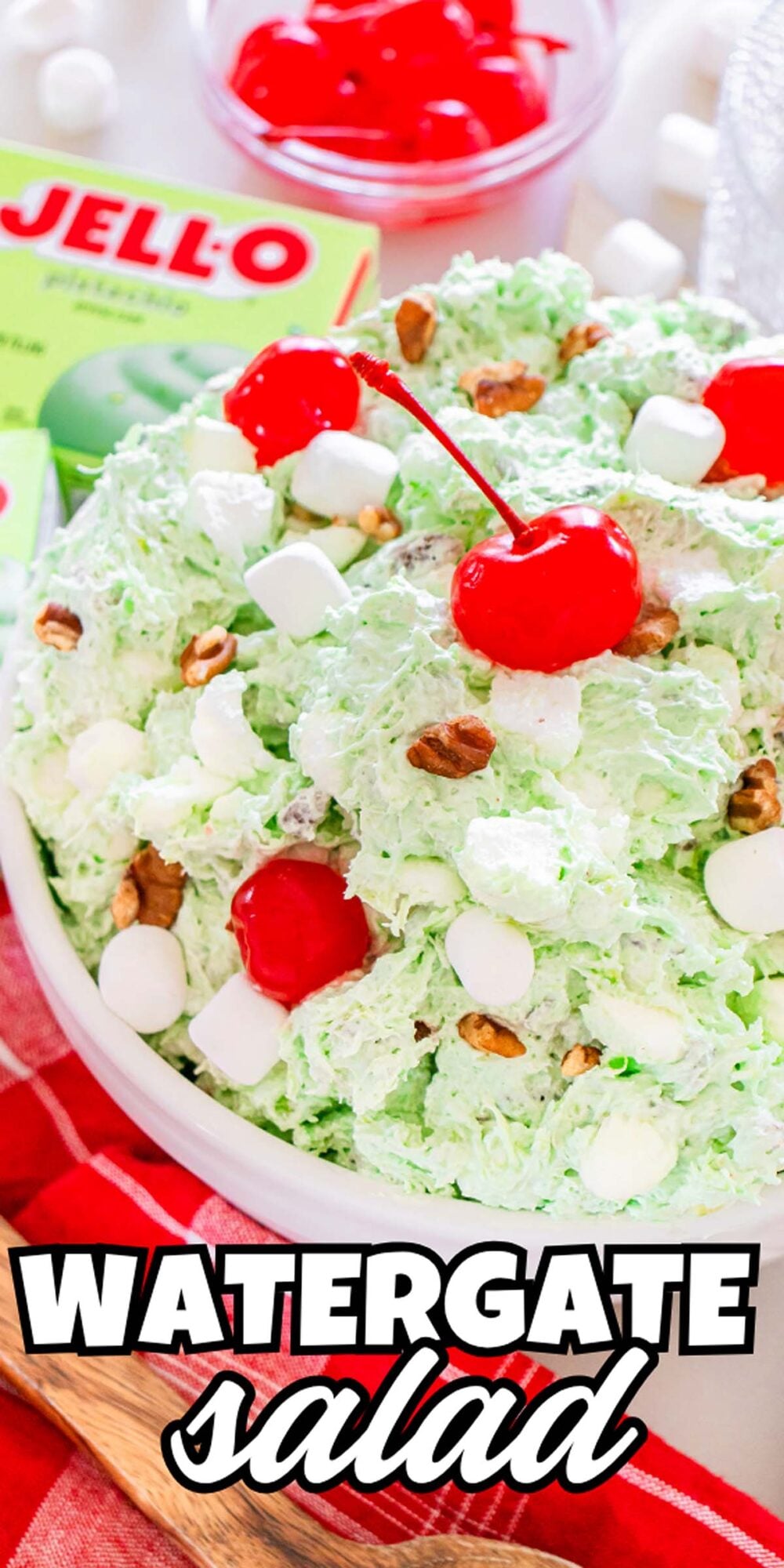 A bowl of classic Watergate Salad topped with cherries, marshmallows, and pecans, with a Jell-O box and extra cherries in the background.