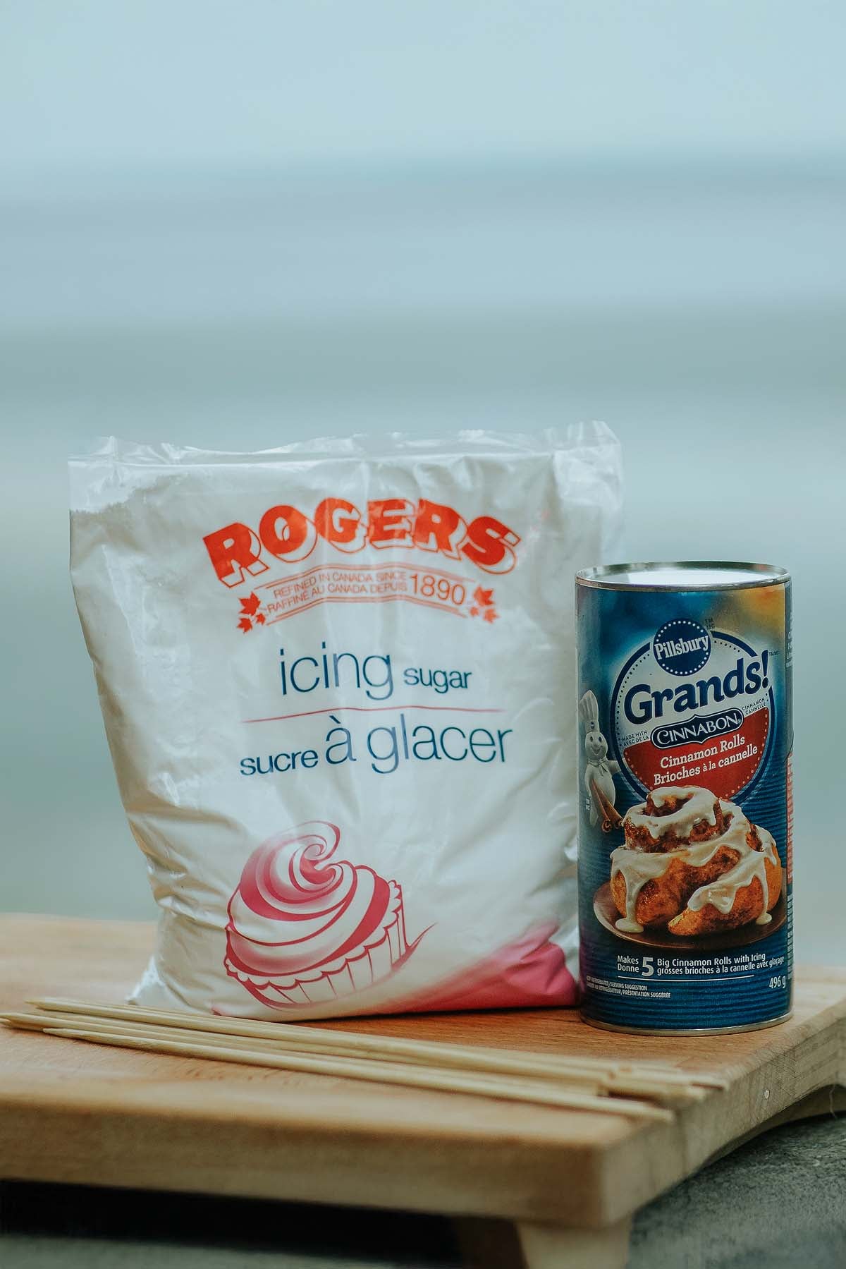 A bag of icing sugar and a can of Pillsbury Grands! cinnamon rolls on a wooden surface&mdash;perfect for making campfire cinnamon twists.